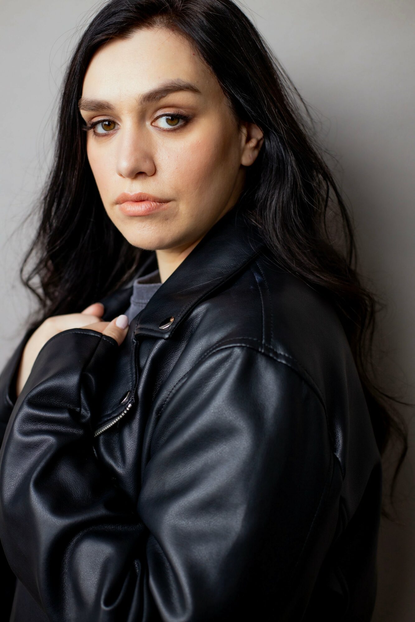 Young woman with long dark hair wearing a black leather jacket, looking at the camera against a gray background.