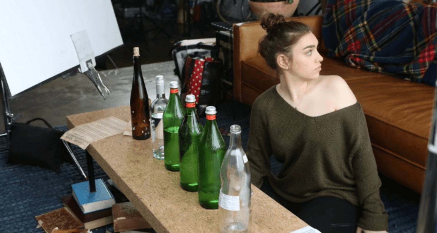 Young woman with brown hair in a bun, wearing an off-shoulder green sweater, sitting on a couch near a table with bottles.