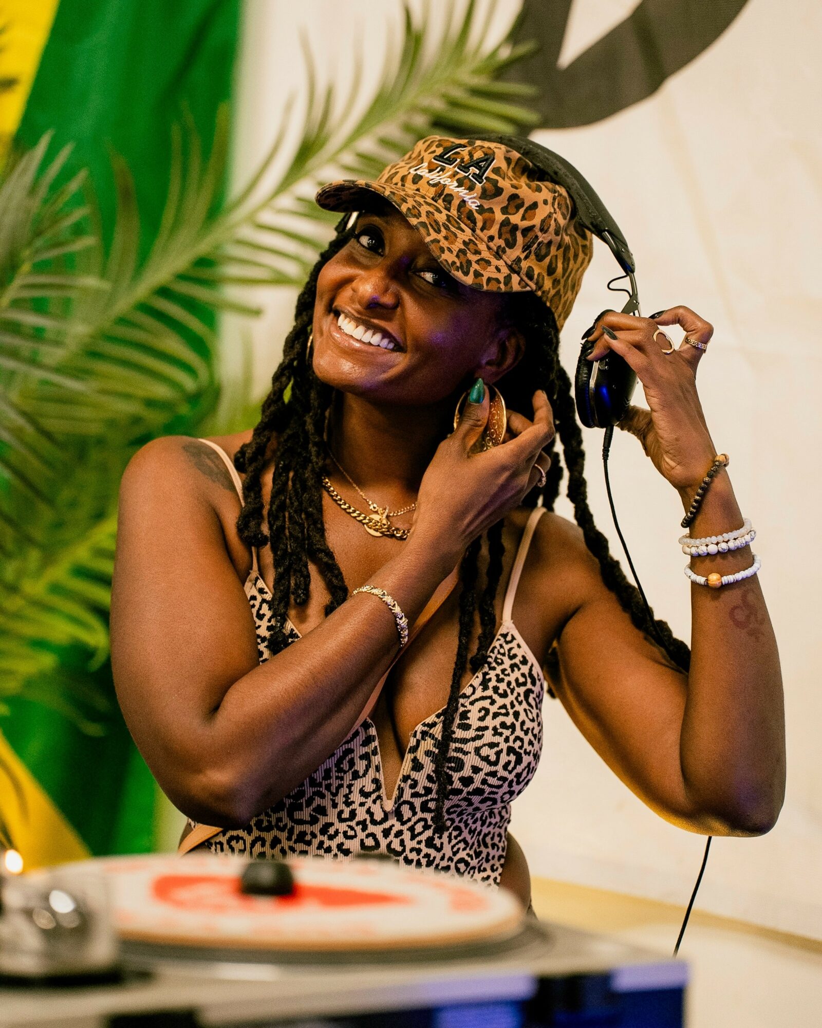 Smiling woman with dreadlocks wearing a leopard print hat and top, listening to music with headphones, surrounded by green plants.