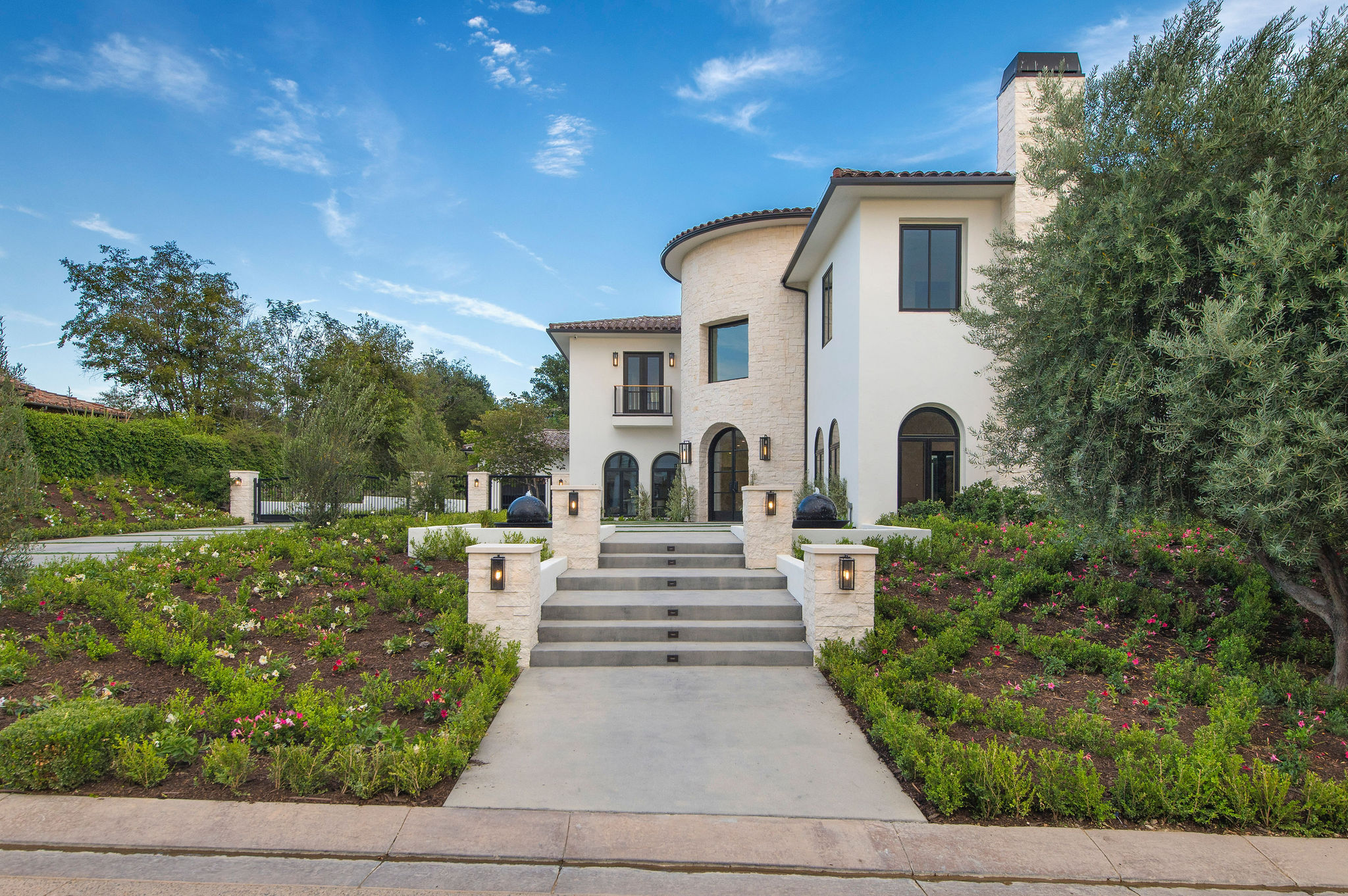 White mansion with curved tower, stairs, surrounded by landscaped garden and trees, under blue sky with clouds.