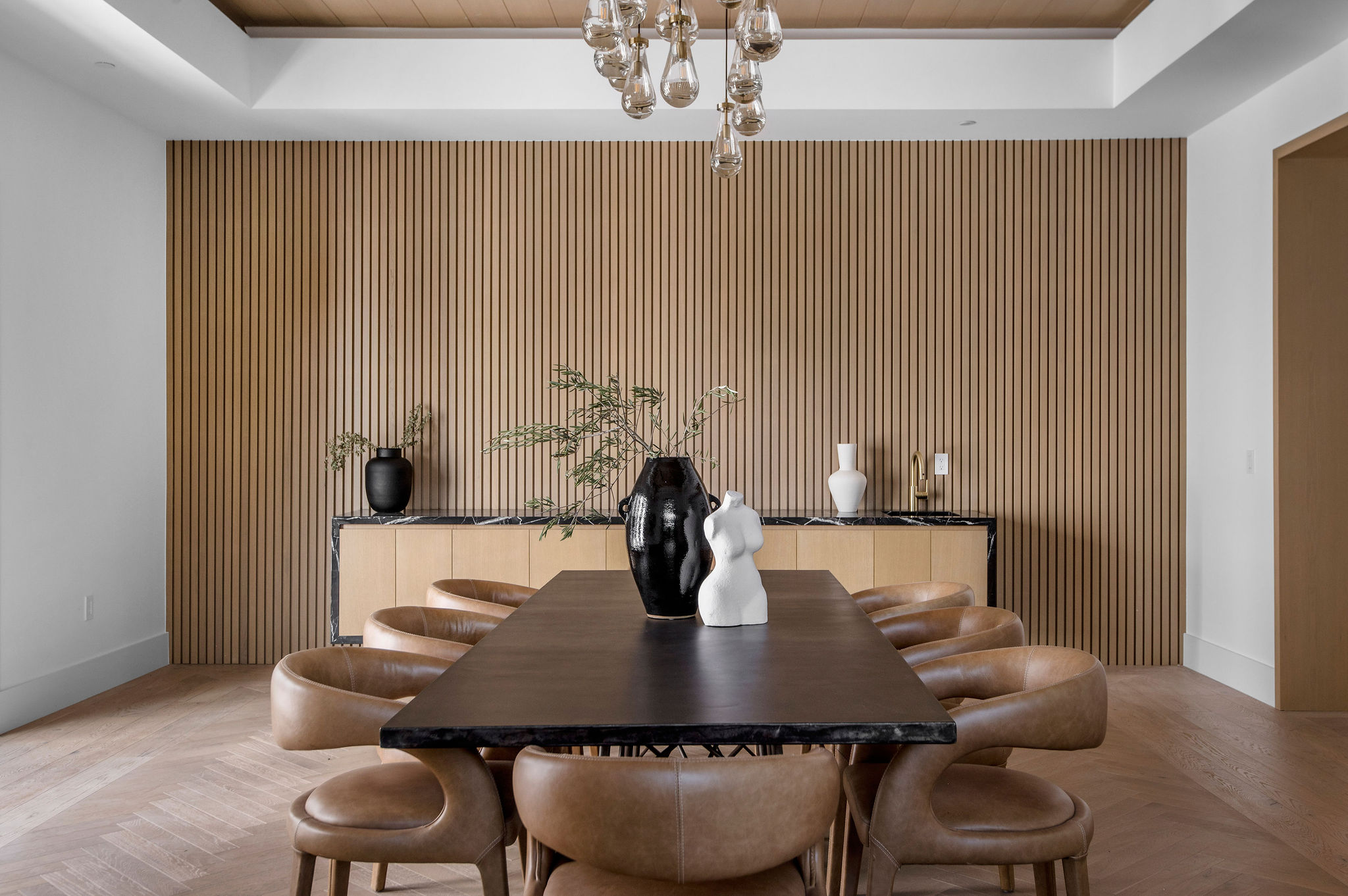 Dining room with wooden wall, dark table, eight beige chairs, and decorative vases and plants on sideboard.