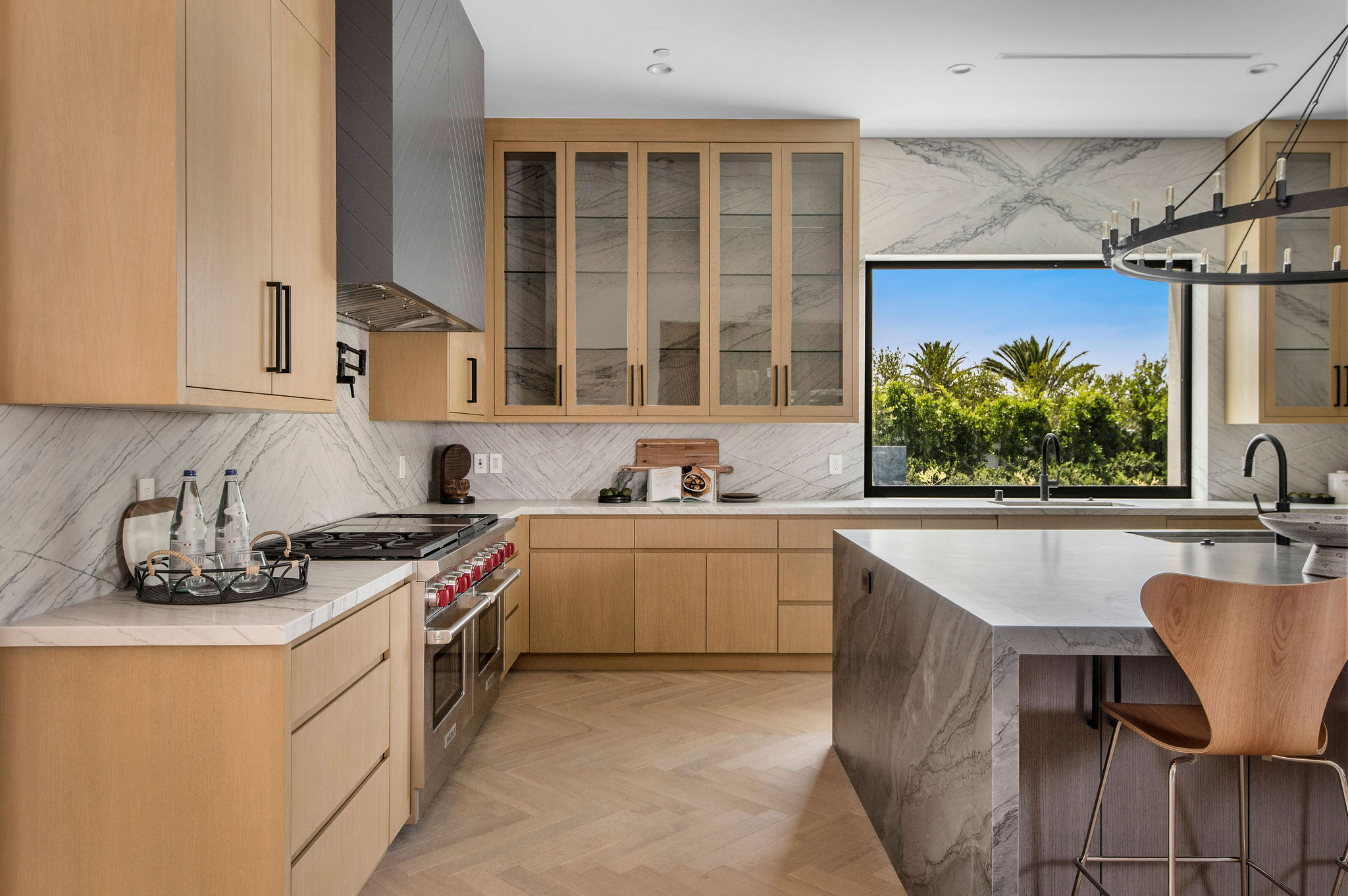 Modern kitchen with wooden cabinets, marble countertops, and a large window showing trees outside.