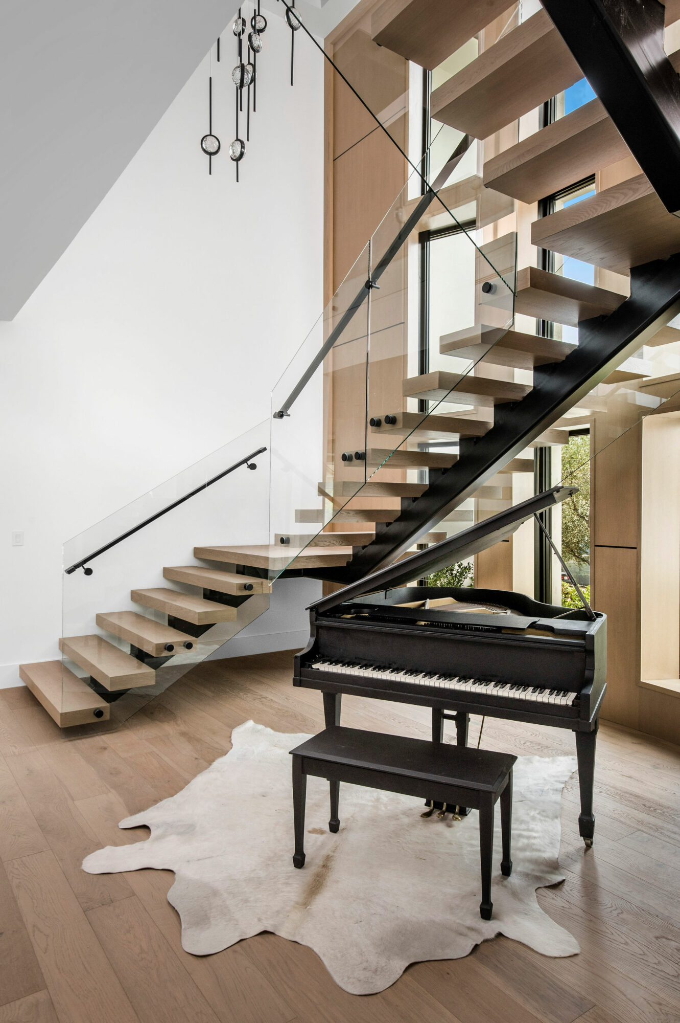 Modern staircase with glass railing and wooden steps, with a black piano and bench on a white rug in a bright room.