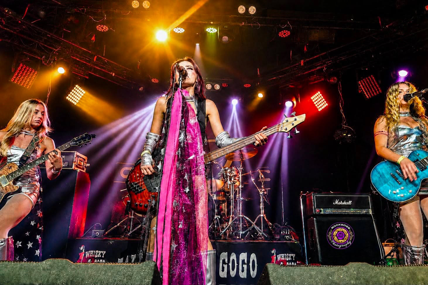Four women perform on stage with colorful lights, one singing into a microphone and playing guitar, others playing instruments.