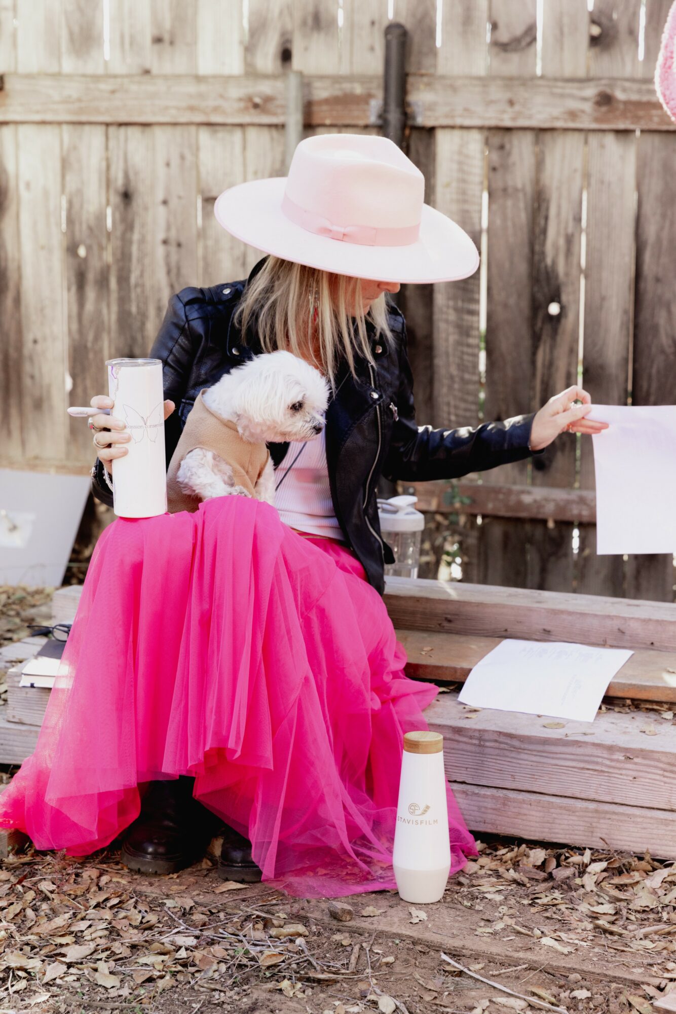 Person wearing a large pink hat and pink skirt sitting on a bench with a small white dog, holding a paper, outdoors.