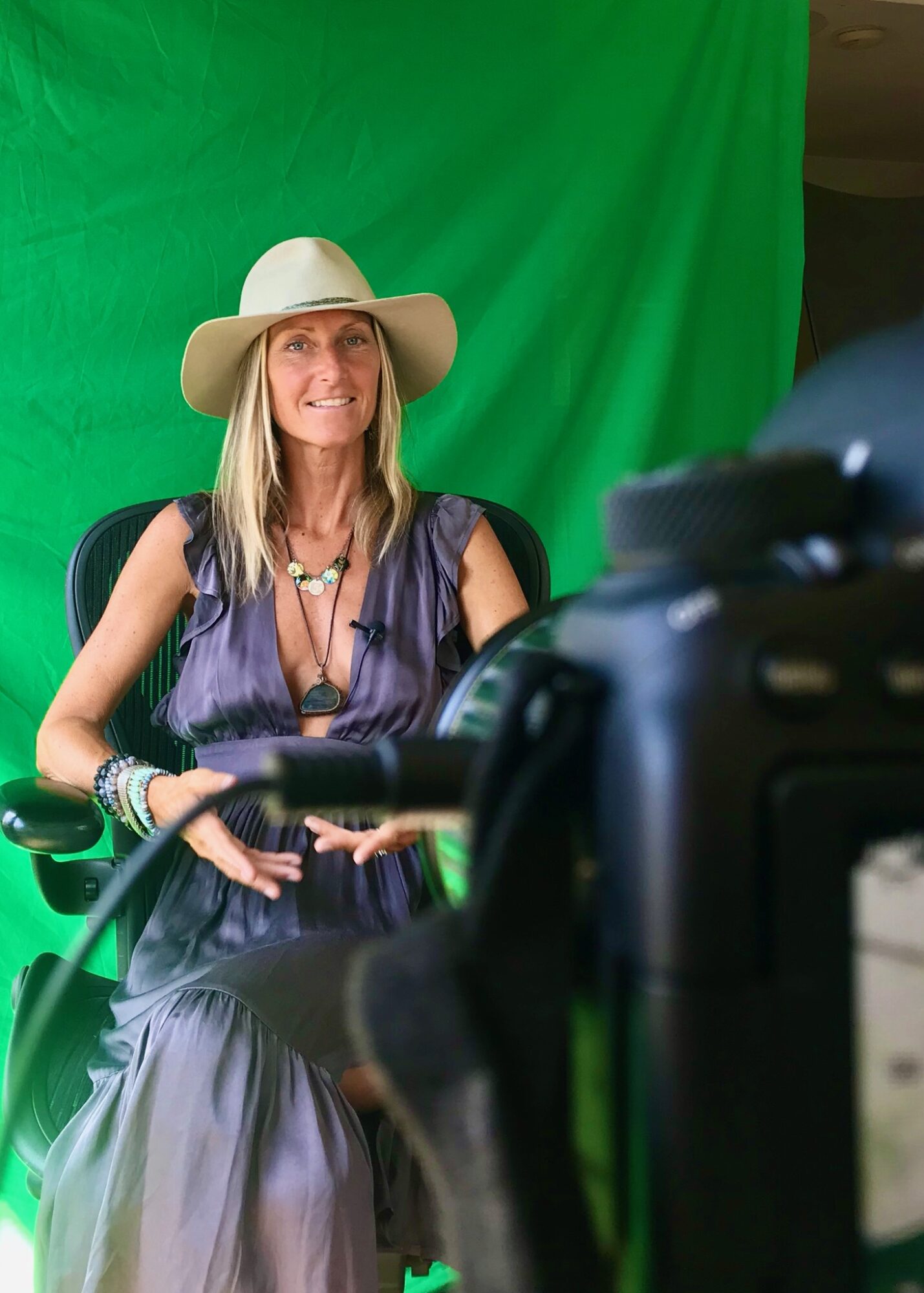 Woman wearing a wide-brimmed hat and jewelry, sitting in front of a green screen, smiling.
