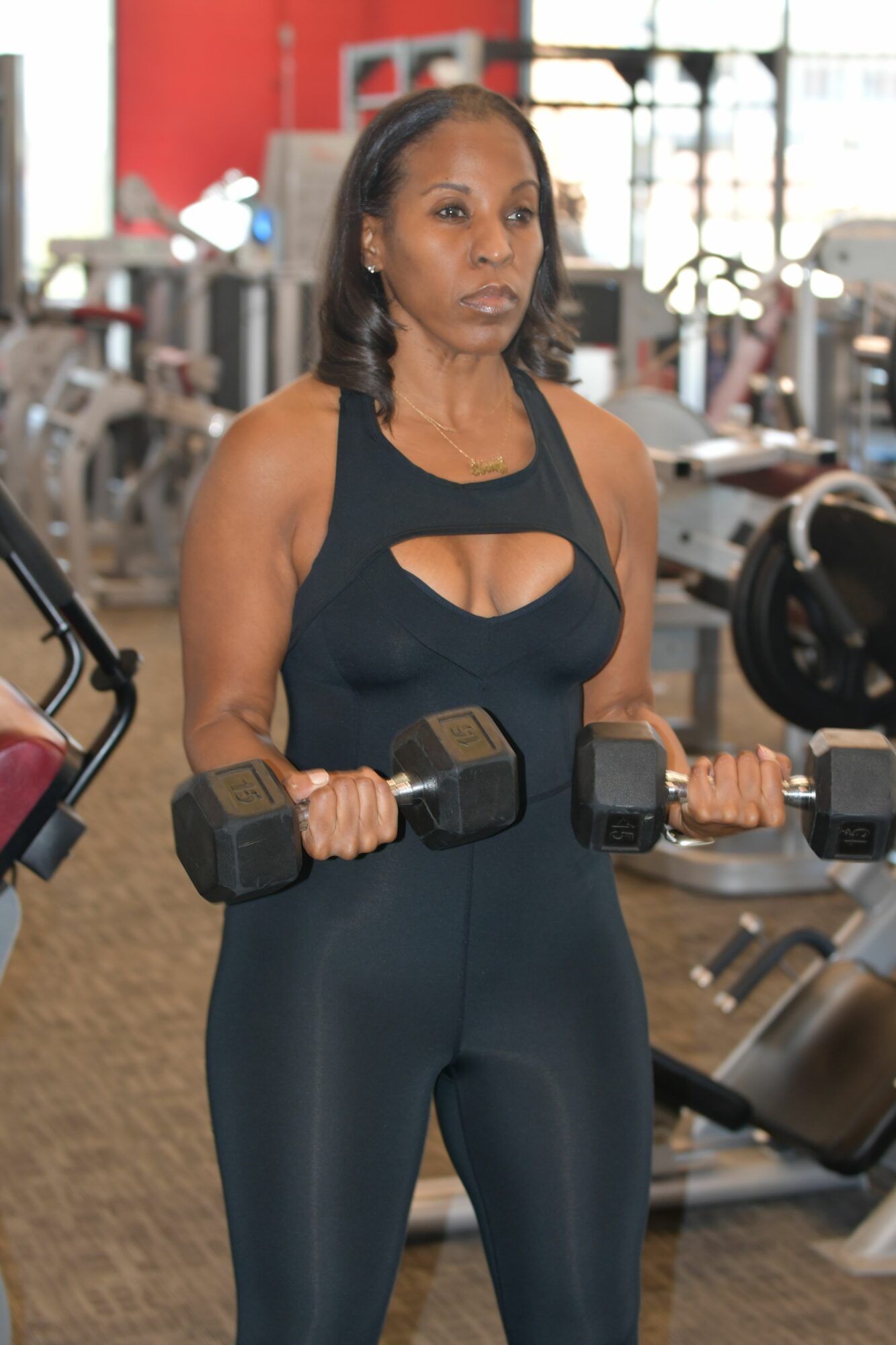 Woman in gym holding dumbbells, wearing black workout outfit, with gym equipment in background.