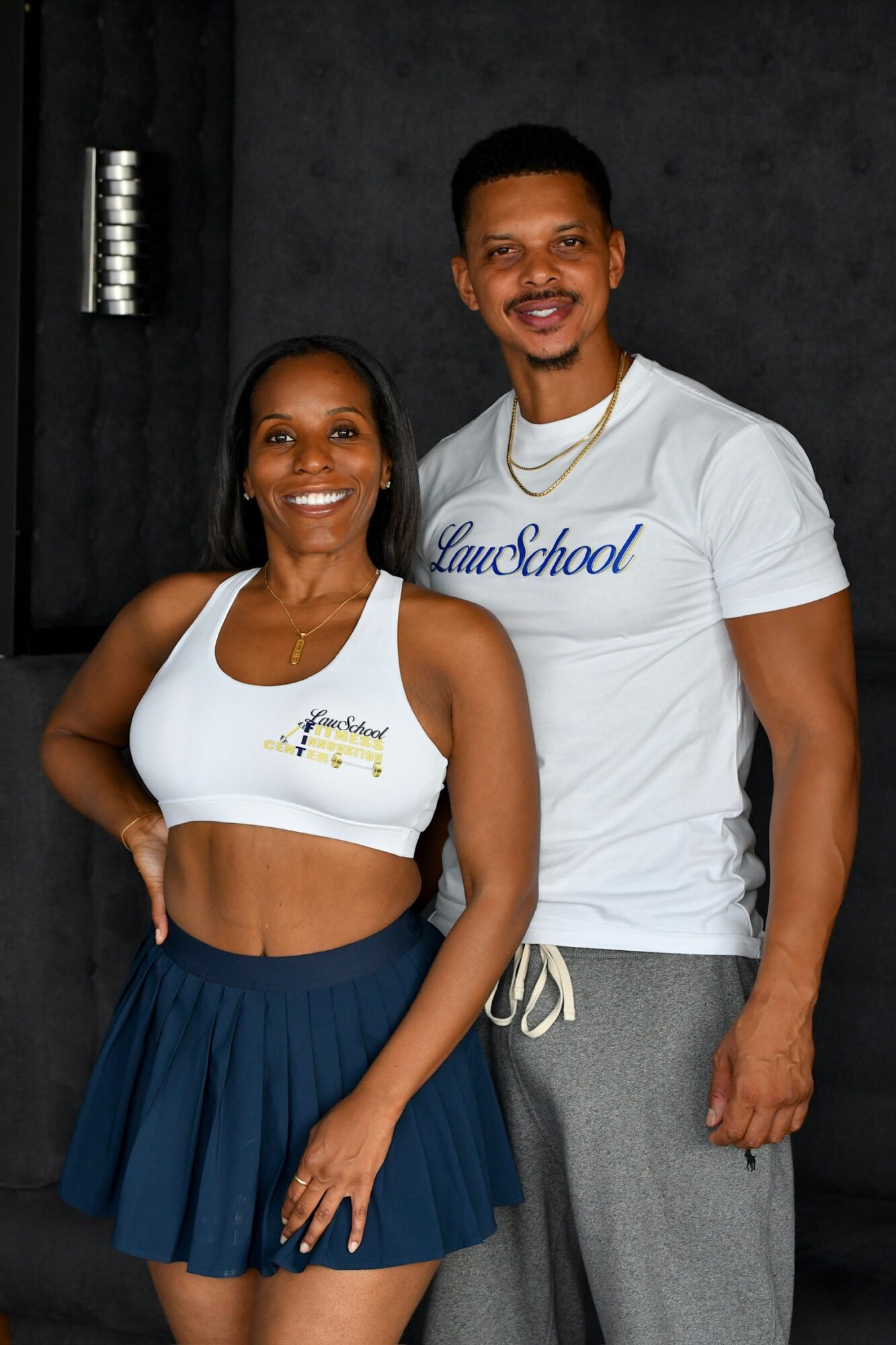 Two smiling people, a woman in a white crop top and a man in a white t-shirt, stand together against a dark background.