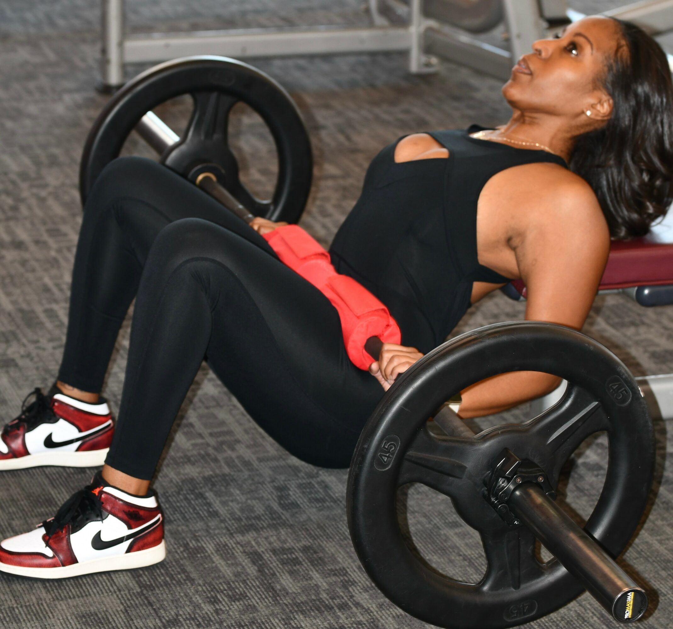 Woman lying on a bench with large wheels, wearing black athletic clothing and sneakers, in a gym setting.