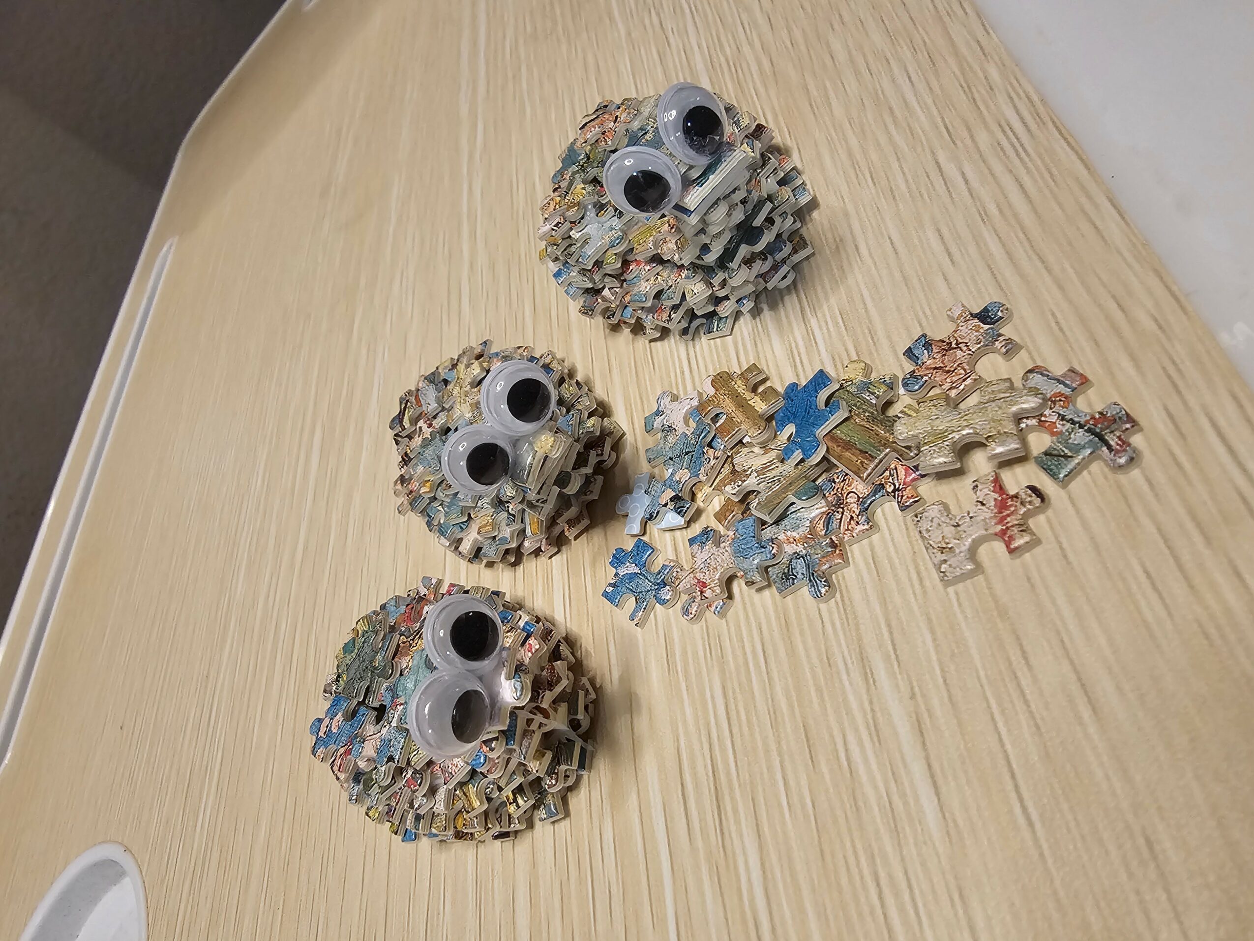 Three owl-shaped objects made from crumpled paper with googly eyes on a light wooden surface, with scattered puzzle pieces nearby.