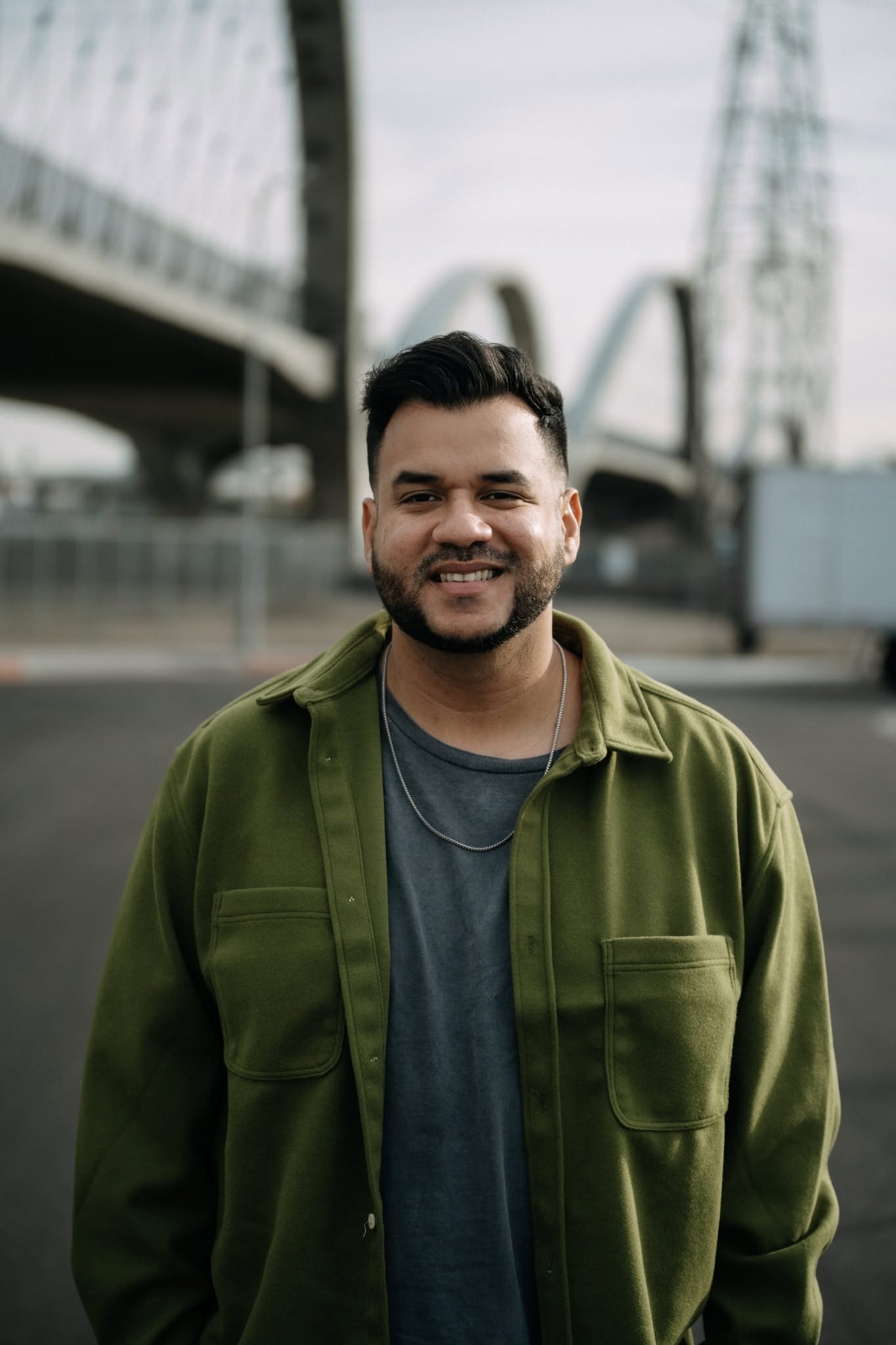 Smiling man with dark hair and beard wearing a green jacket and gray shirt outdoors, cityscape background.