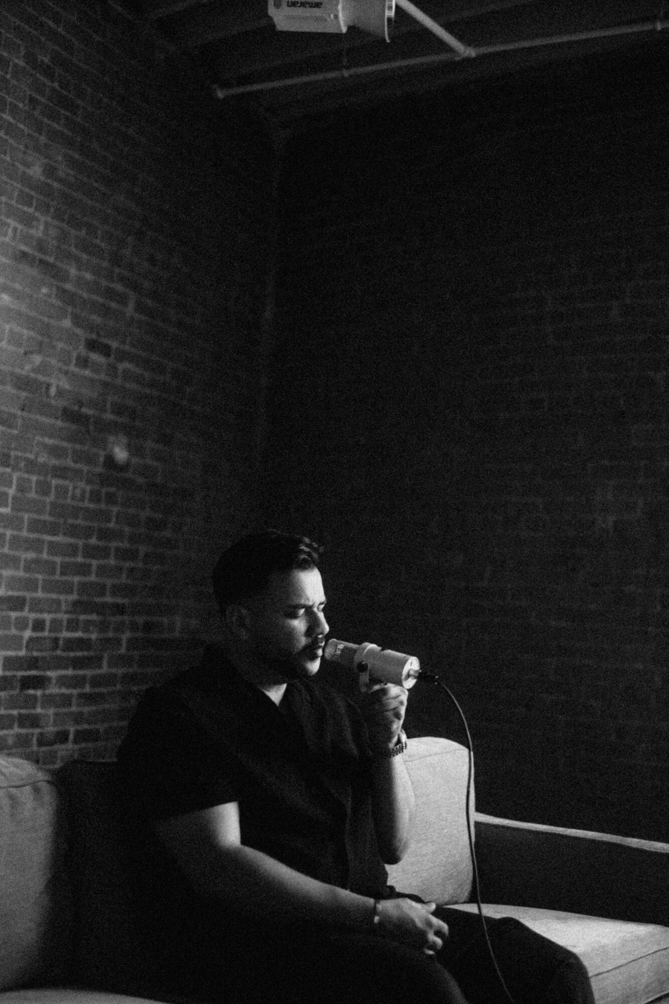 Man with a beard sitting on a couch, holding a microphone, against a brick wall background.