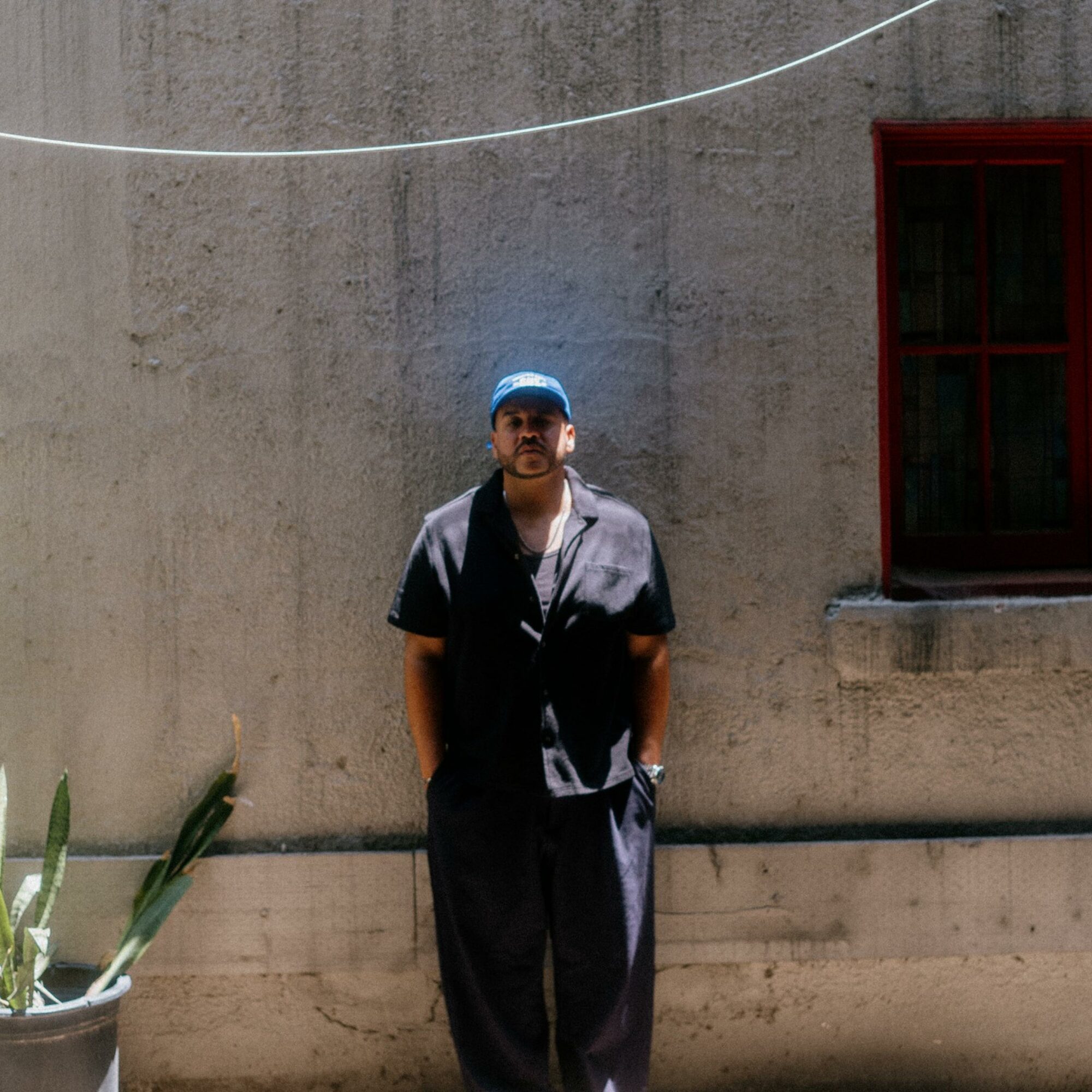 Man standing against a concrete wall with a window, wearing a cap and casual clothing.