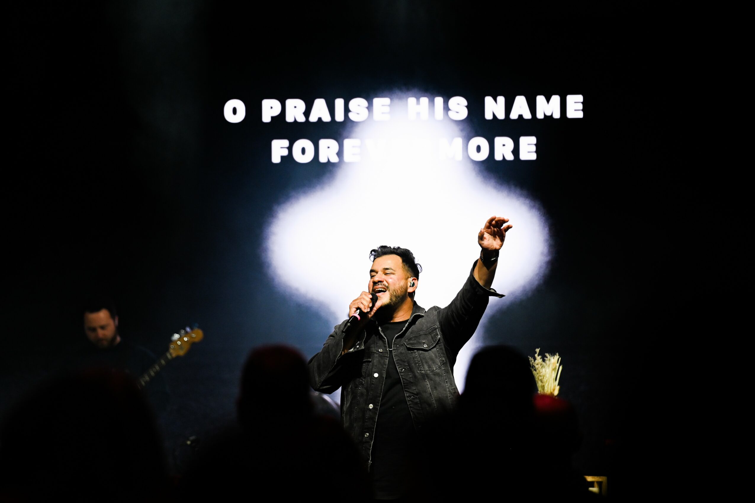 A man singing on stage with a microphone, raising his hand, with a bright light behind him and text on screen.