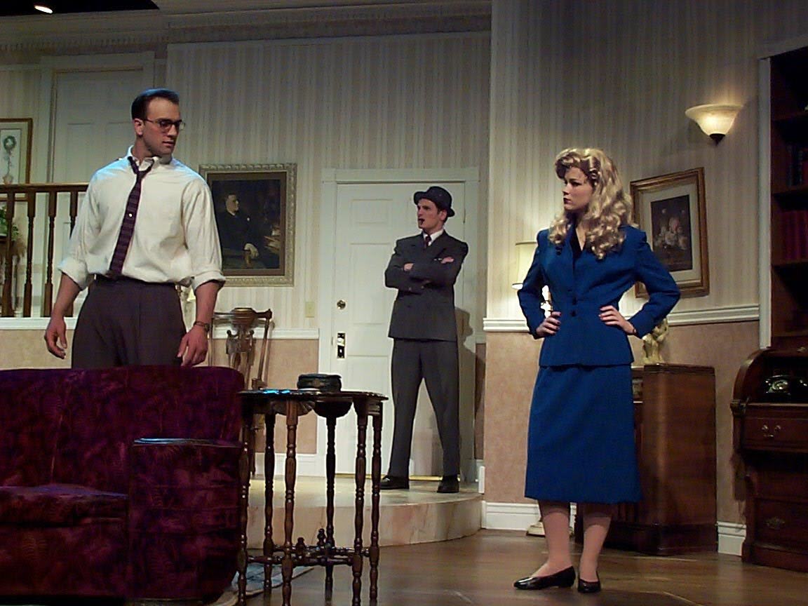Three actors on a stage set resembling a living room, with furniture and framed pictures, engaged in a scene.