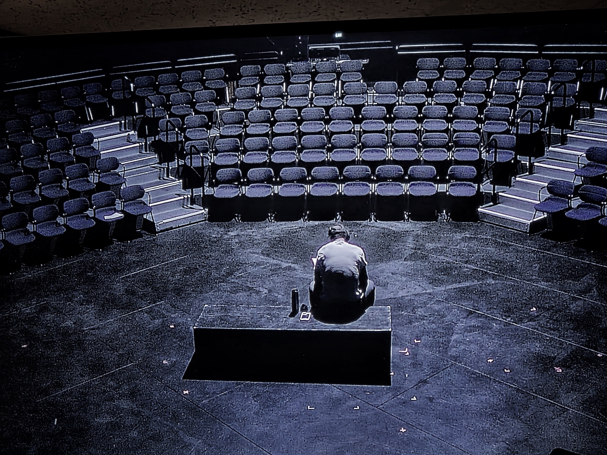 A person sitting alone on a stage in an empty theater with many rows of seats surrounding them.