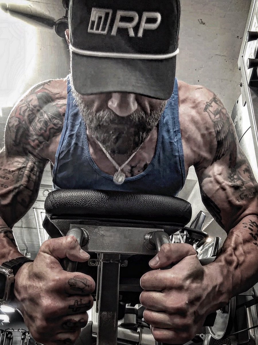 Man with tattoos wearing a cap and tank top, lifting weights in a gym, focusing on his arms and upper body.
