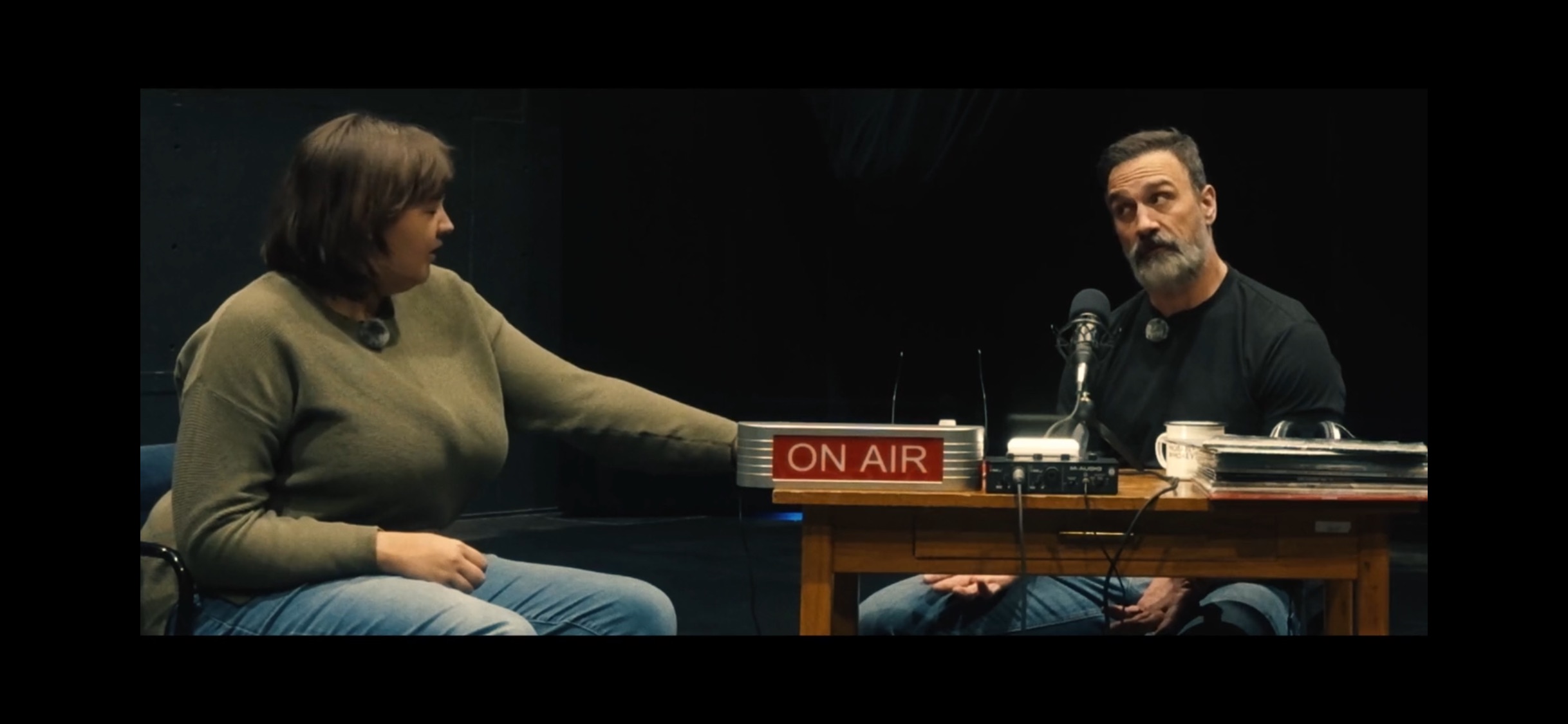 Two people sit at a table with a microphone, a sign reads 'ON AIR,' in a dark studio setting.