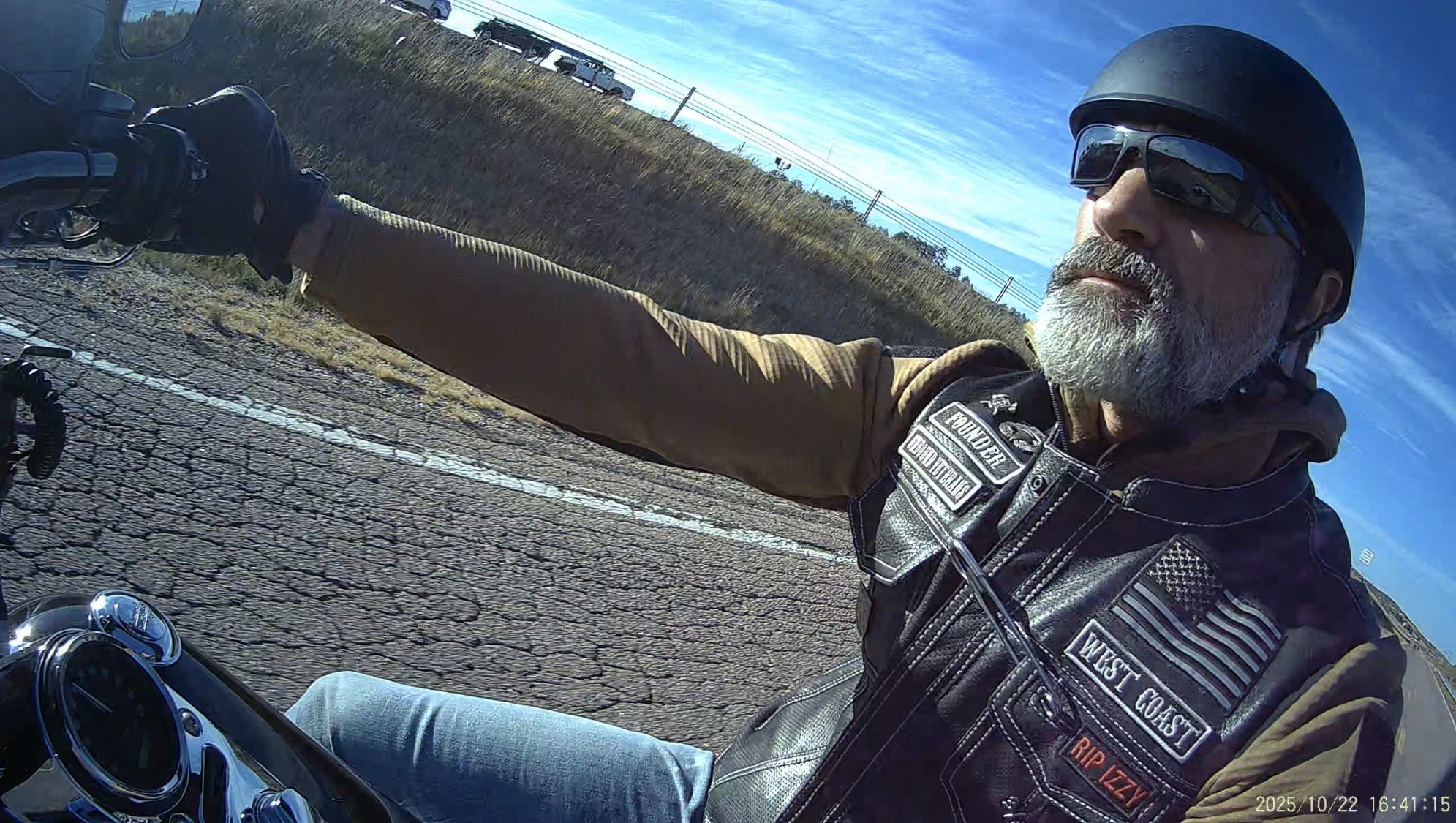 Motorcycle rider with sunglasses and helmet extending arm, outdoors on a sunny day, background of open sky and landscape.