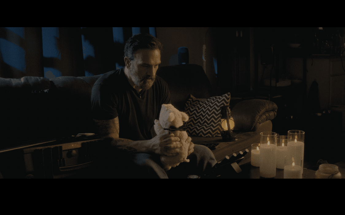 Man sitting on a couch in a dimly lit room, holding a cloth, with candles on a table nearby.
