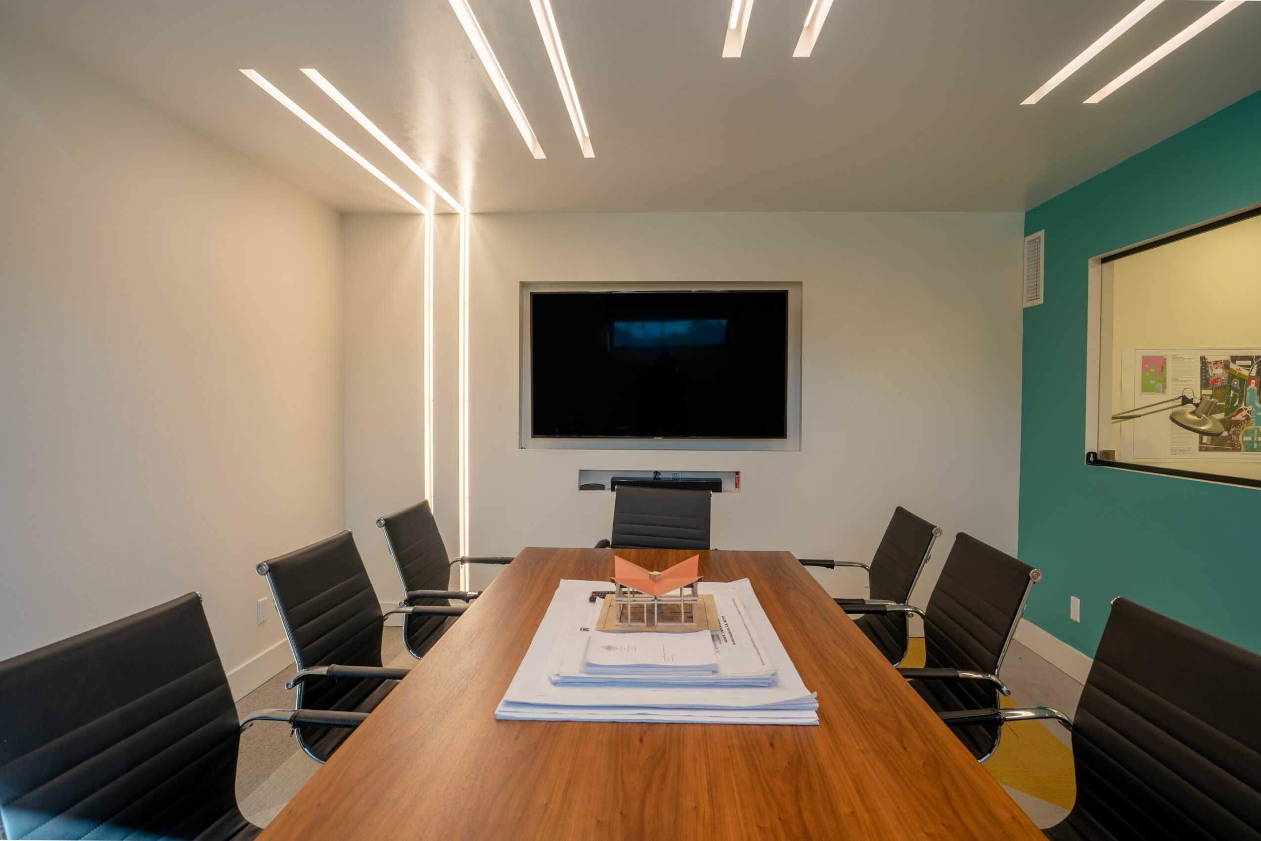 Conference room with a wooden table, six black chairs, a wall-mounted TV, and a whiteboard, illuminated by ceiling lights.