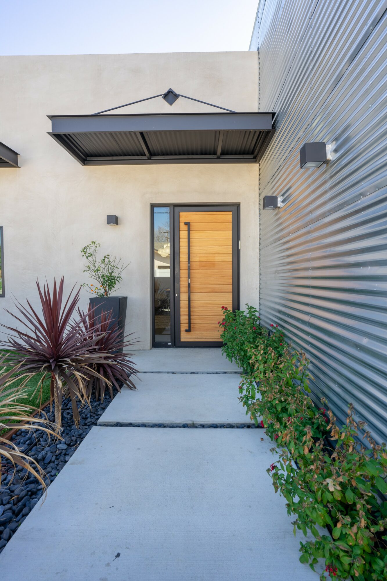 Front entrance with a wooden door, concrete pathway, plants on both sides, and modern exterior walls.