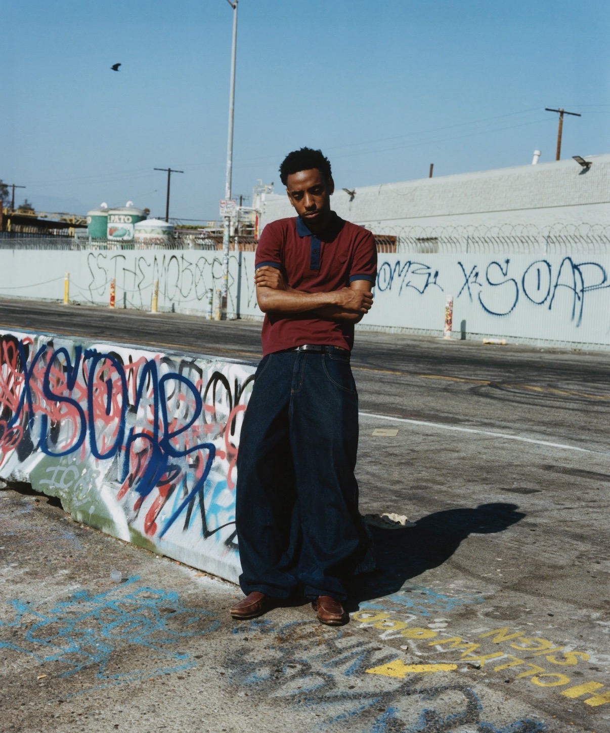 Person standing with arms crossed near graffiti-covered wall, sunny day, urban setting, clear blue sky.