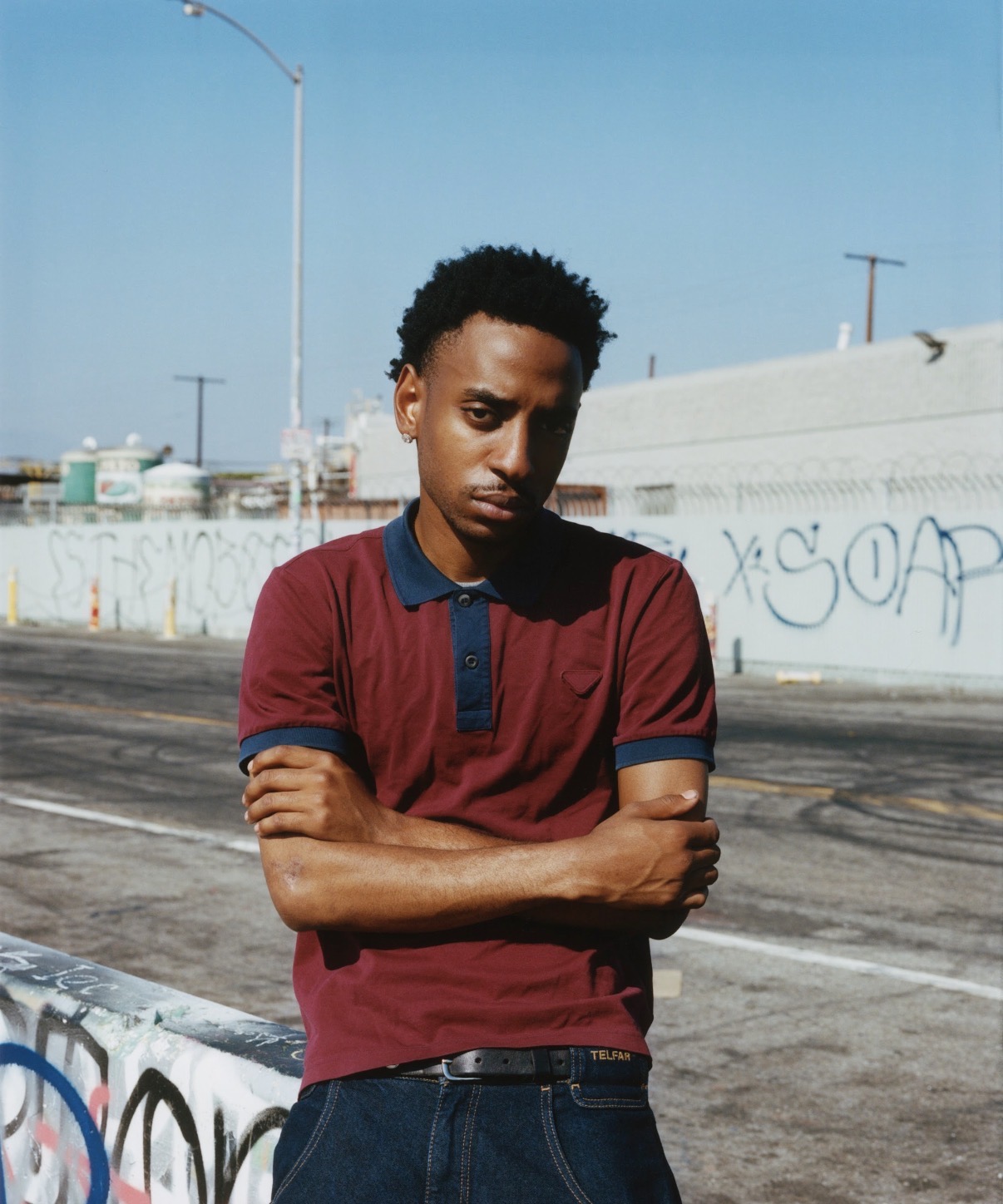 Young man with crossed arms standing outdoors on a street with graffiti wall behind him, under a clear blue sky.