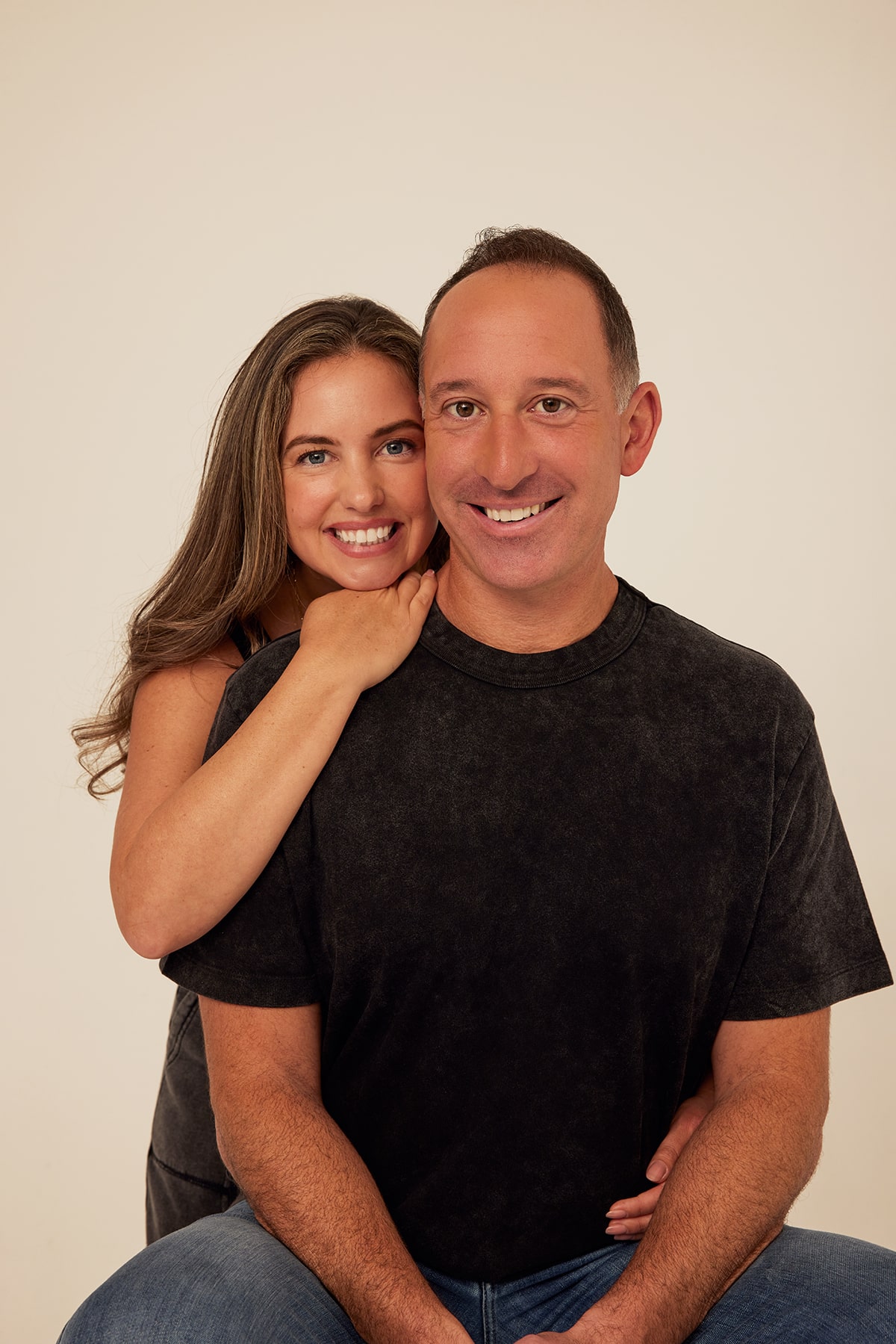 A smiling woman with long hair leans on a smiling man with short hair, both facing the camera against a plain background.