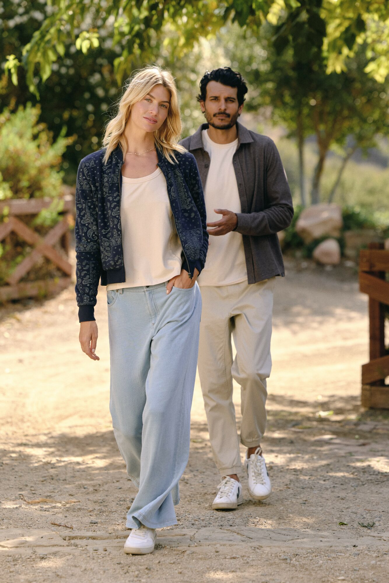 Two people walking outdoors on a sunny day, surrounded by trees and rocks, with a wooden fence in the background.