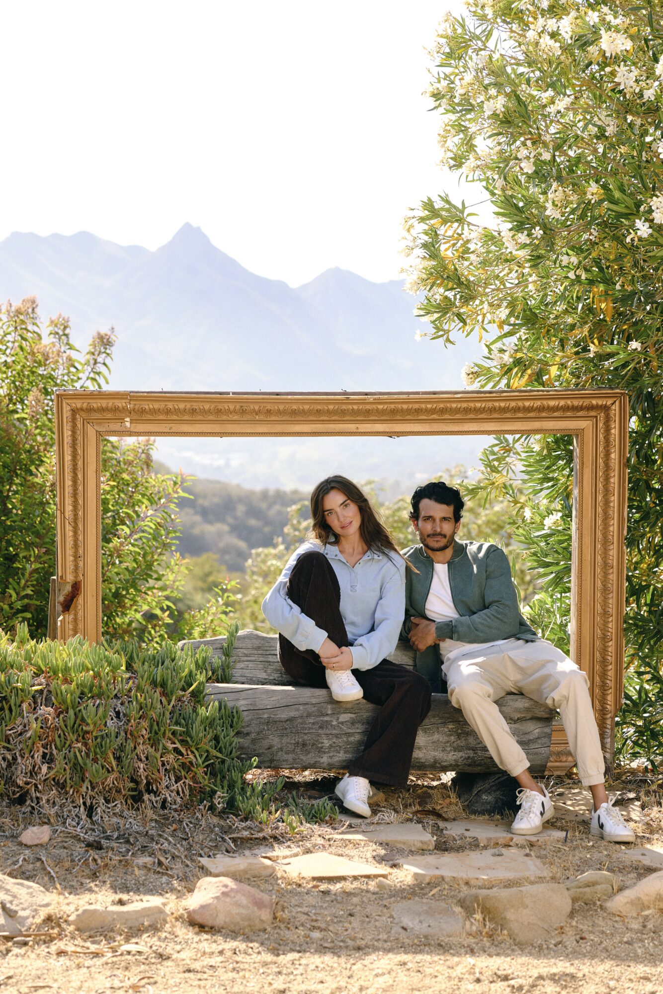 Two people sitting on a bench outdoors, framed by a large wooden frame, with trees and mountains in the background.