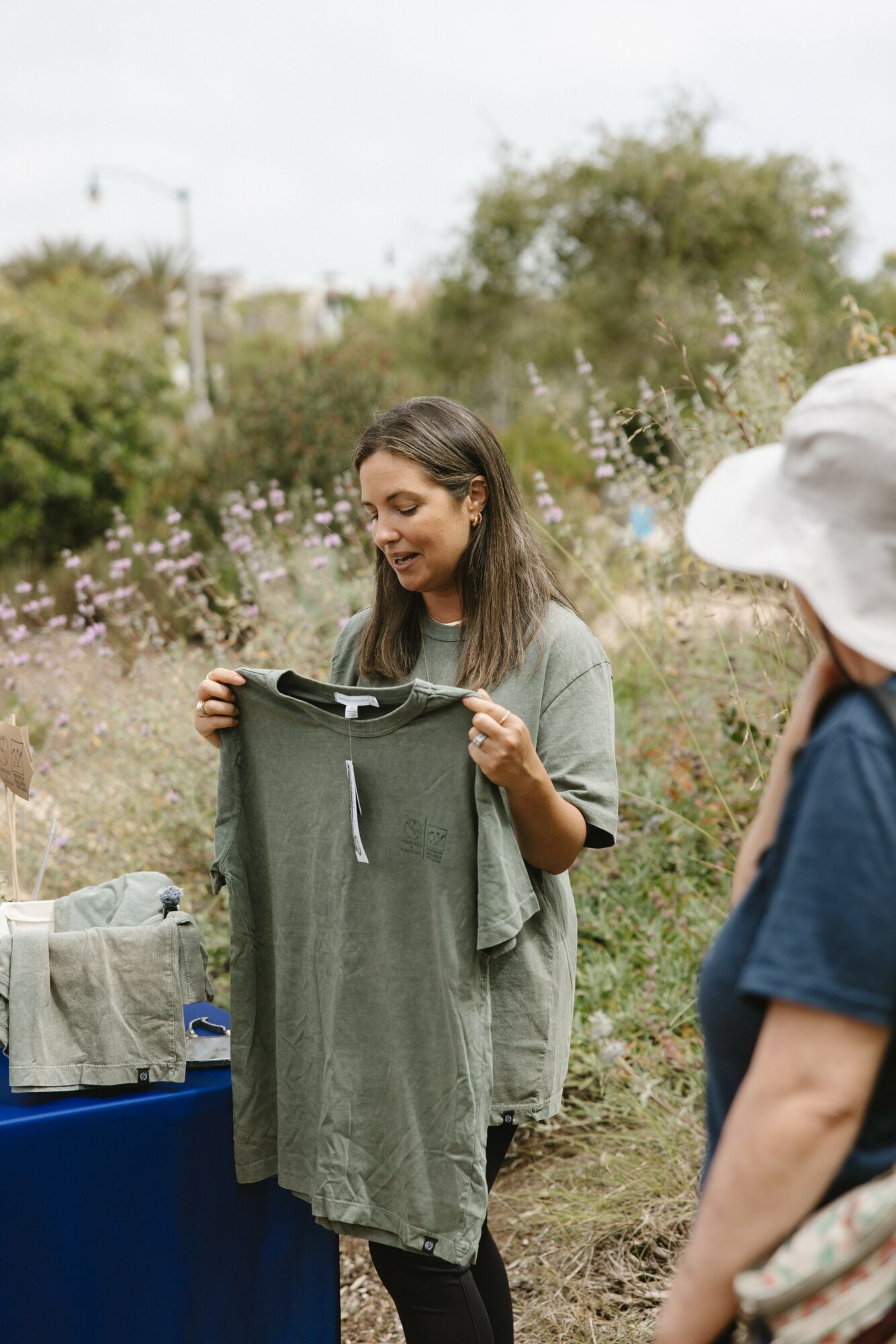 Young woman holding a gray T-shirt outdoors with plants and trees in the background.