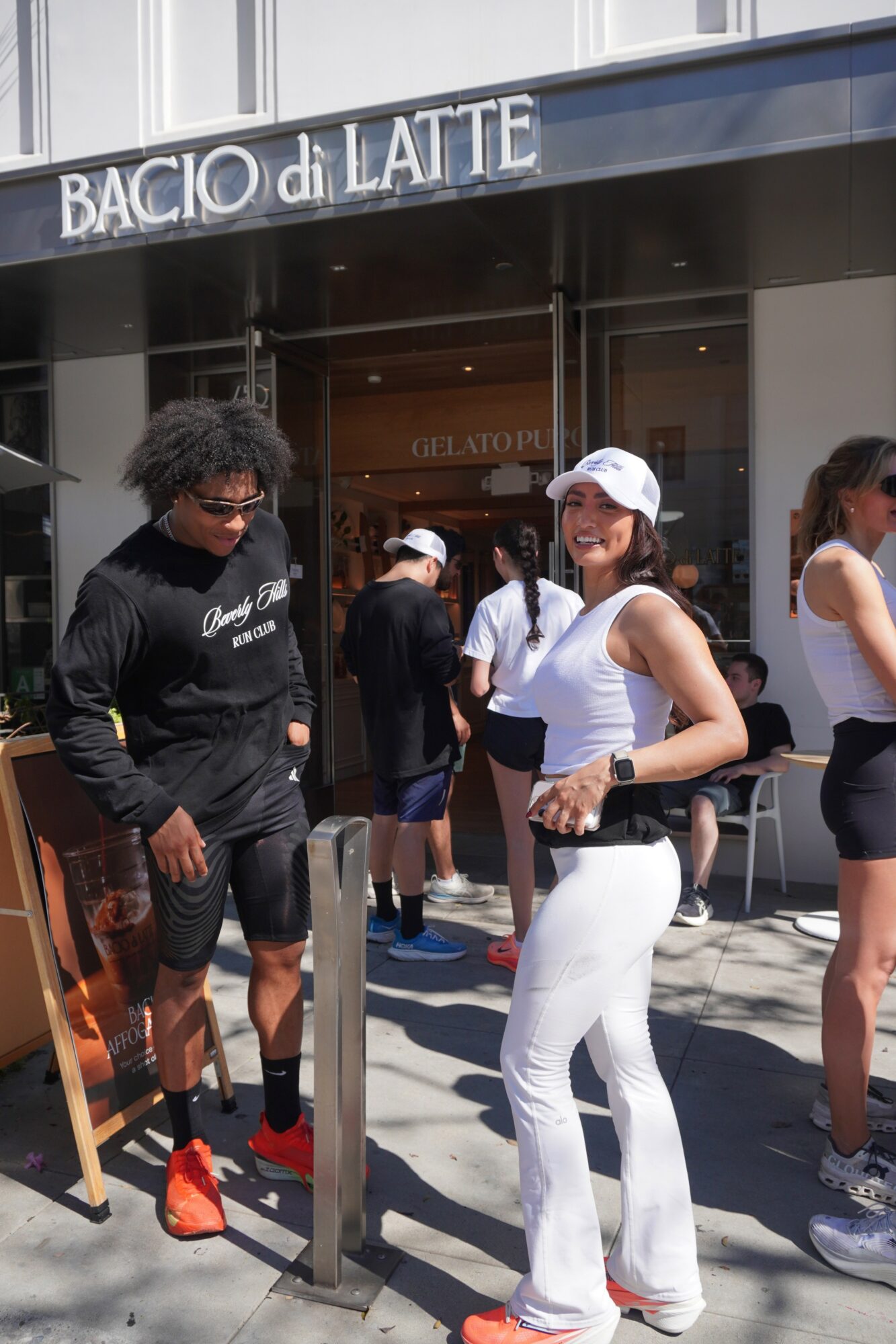 Group of people standing outside a cafe with a sign reading 'BACIO di LATTE', some wearing sunglasses and casual clothing.