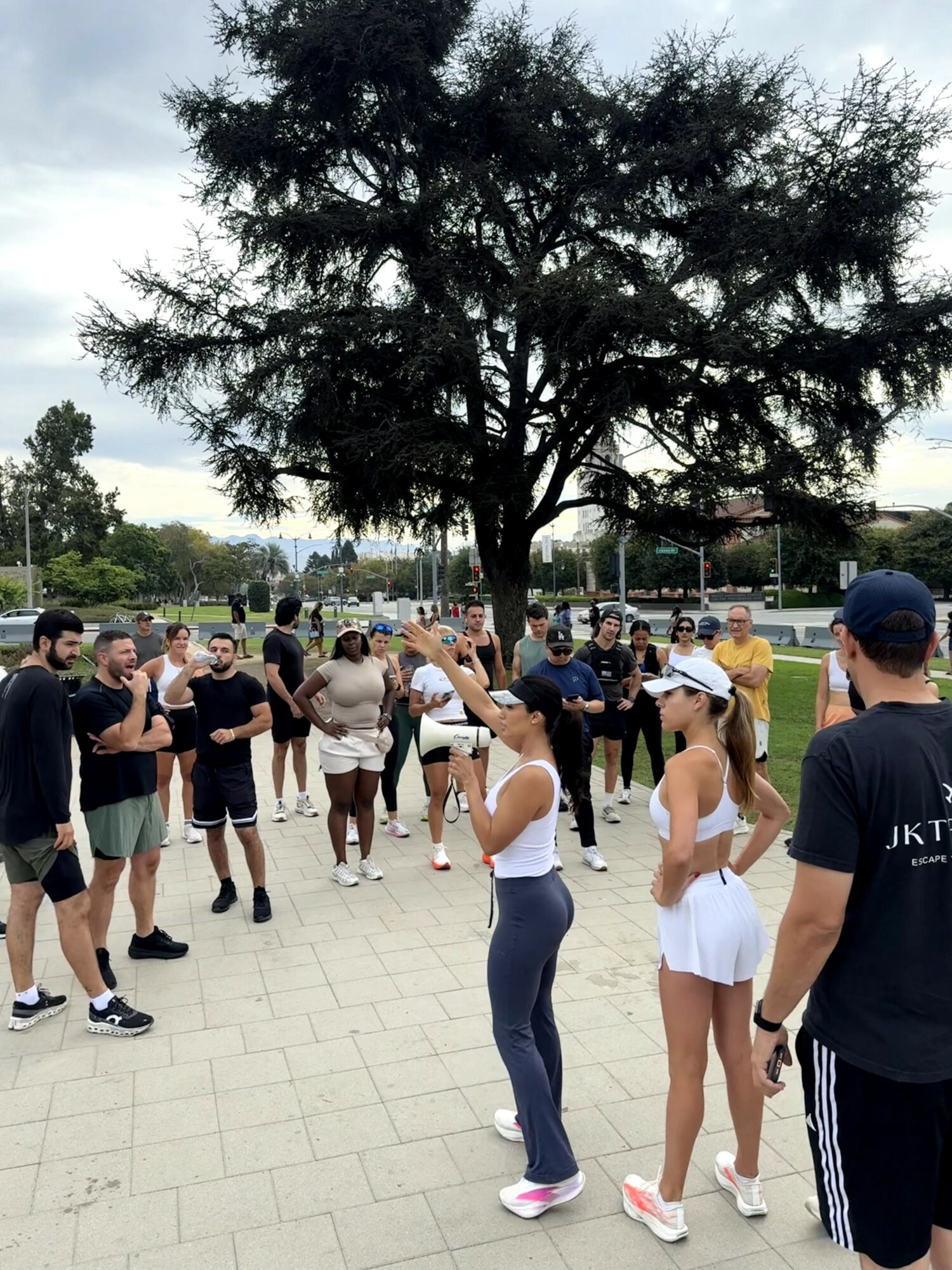 Group of people gathered outdoors near a large tree, some talking and others listening, on a paved area.