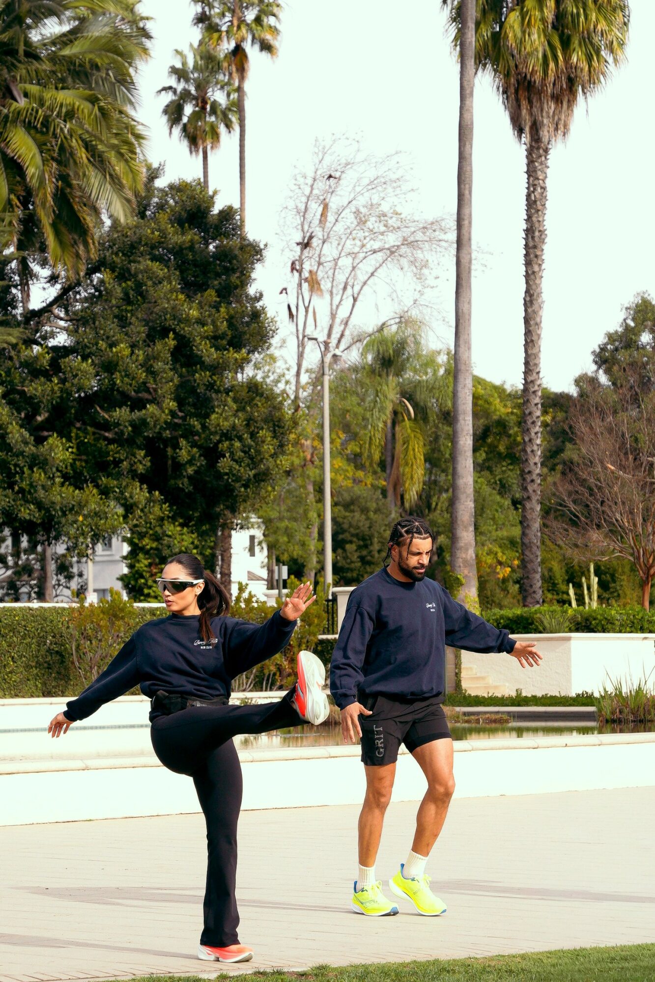 Two people exercising outdoors with palm trees and greenery in the background, one person stretching and the other walking.