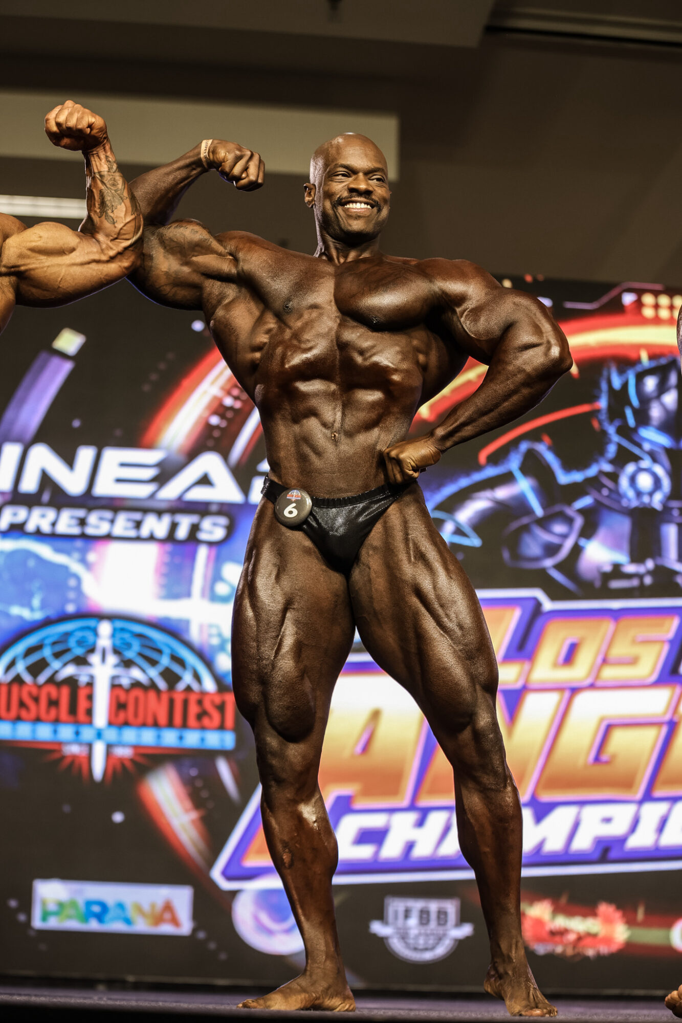 Male bodybuilder on stage flexing muscles, smiling, with a colorful event backdrop, wearing a black posing suit.