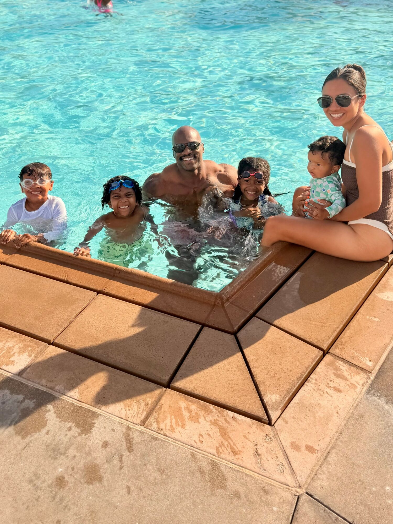 Group of children and an adult sitting in a swimming pool with a woman sitting on pool edge, all smiling and wearing sunglasses.
