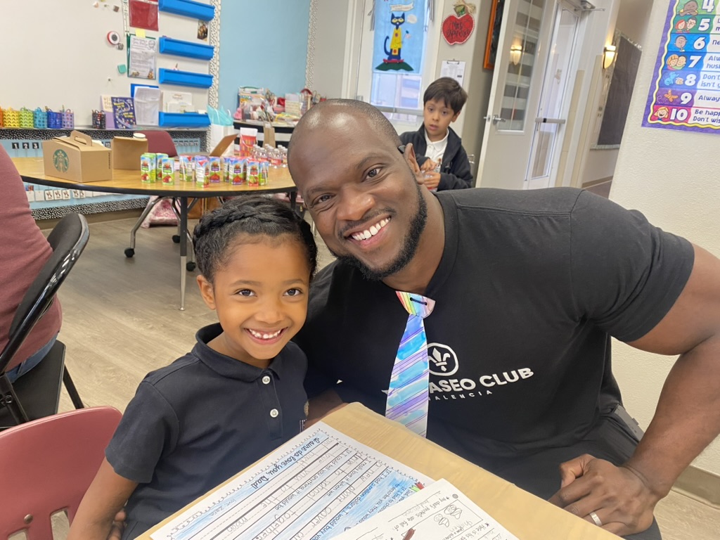 Smiling man and young boy sitting at a table in a classroom, with colorful educational materials in the background.