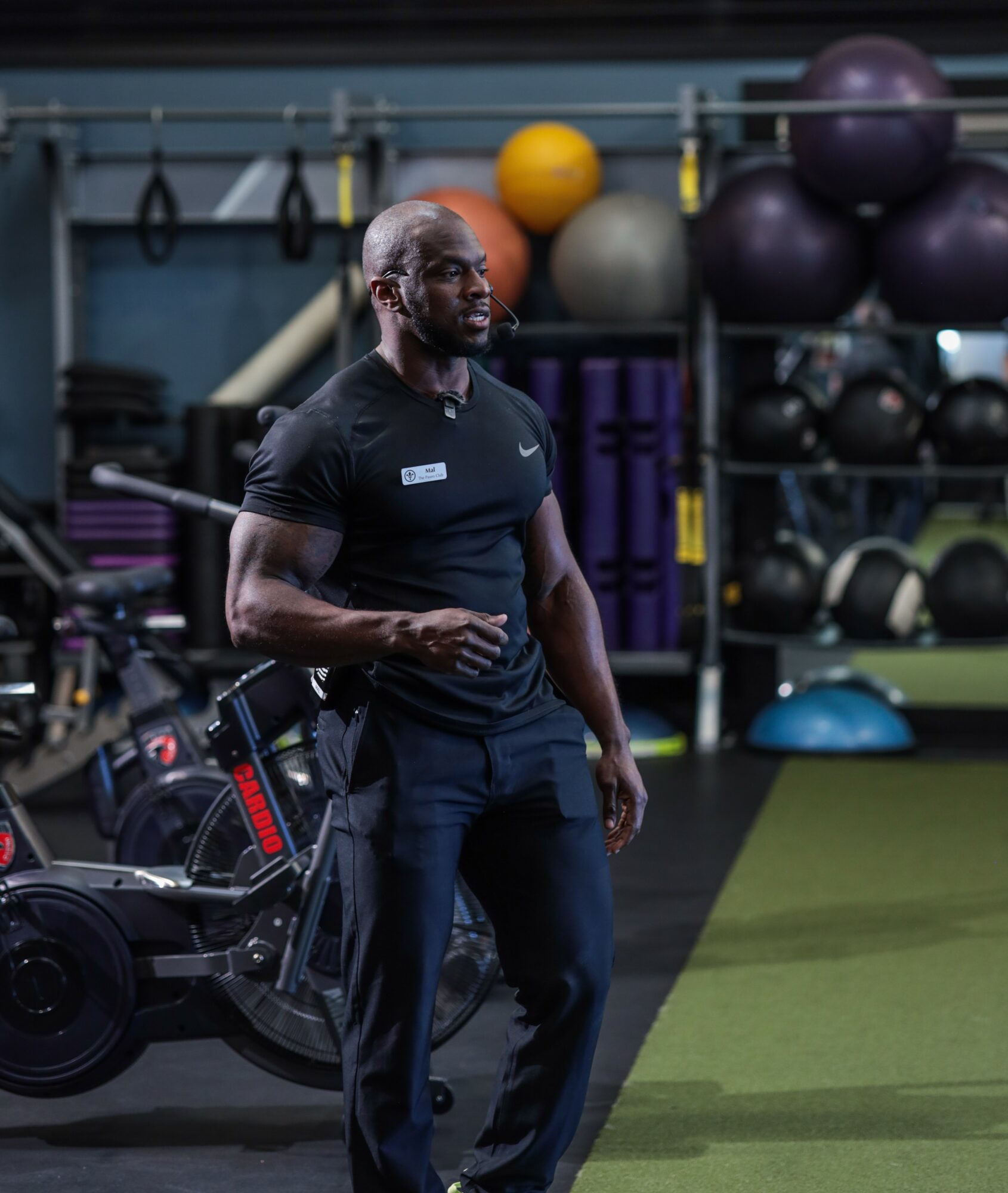 Man in black athletic clothing standing in gym with exercise equipment and large exercise balls in background.