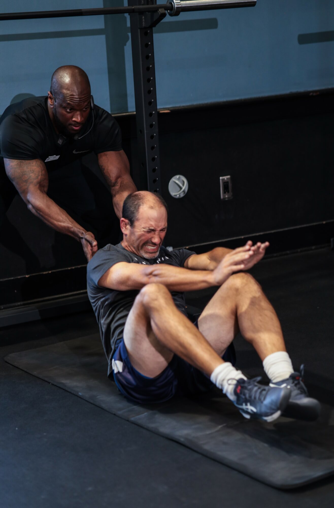 Two men exercising on a mat; one is sitting and smiling, the other is standing behind him, assisting.