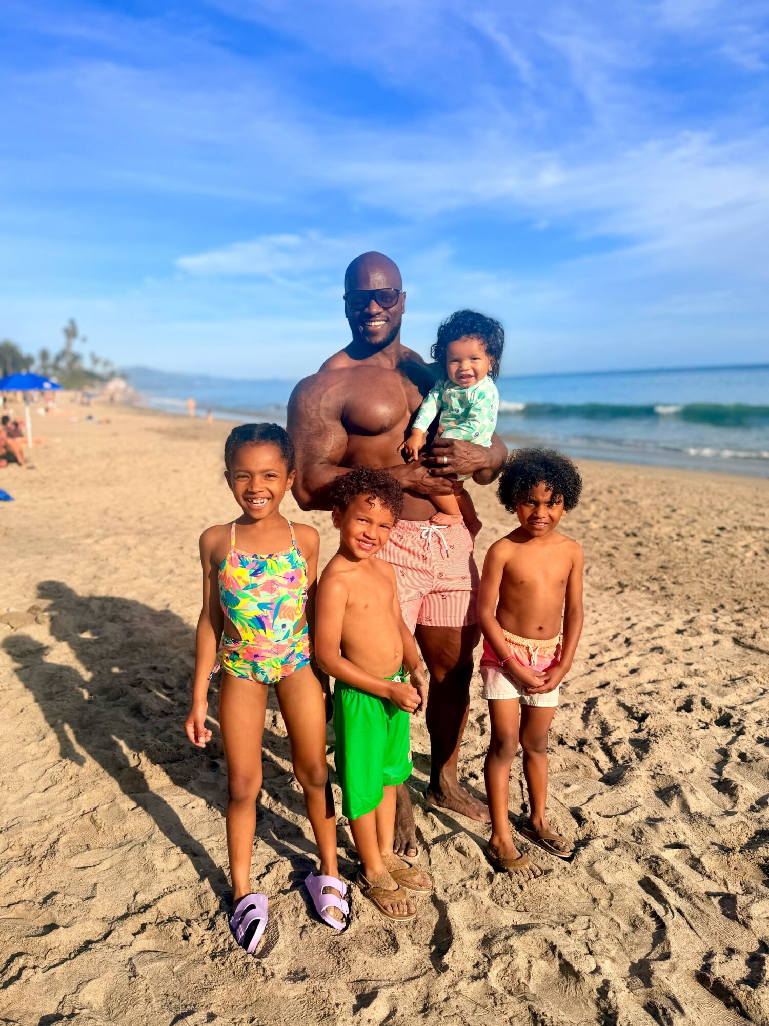 A man with three children on a sandy beach under a blue sky, one child in his arms, others standing nearby.