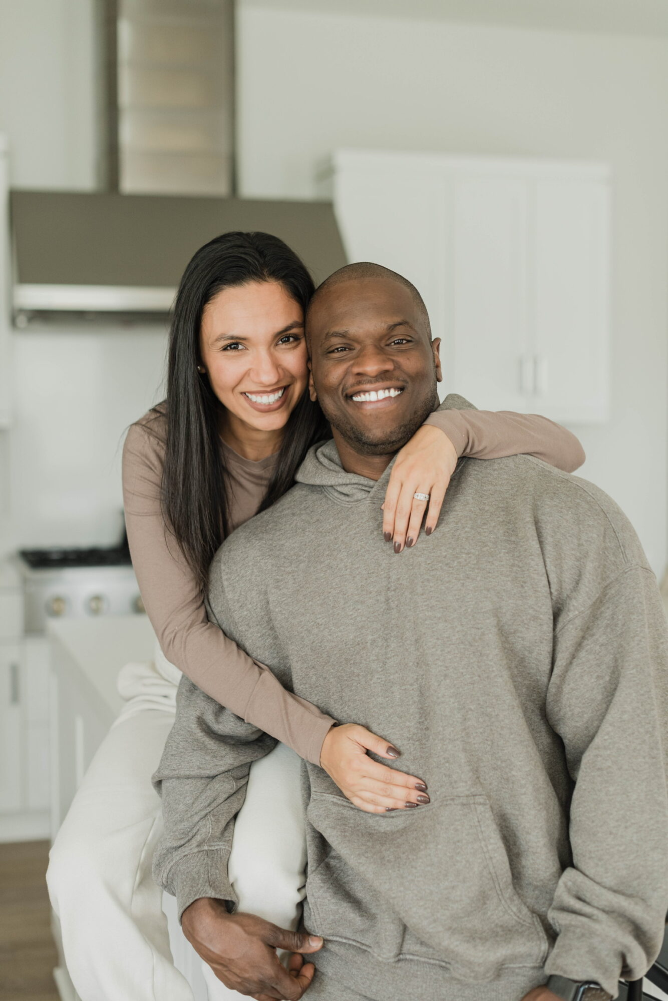 A smiling woman with long dark hair hugging a smiling man with a shaved head in a kitchen.