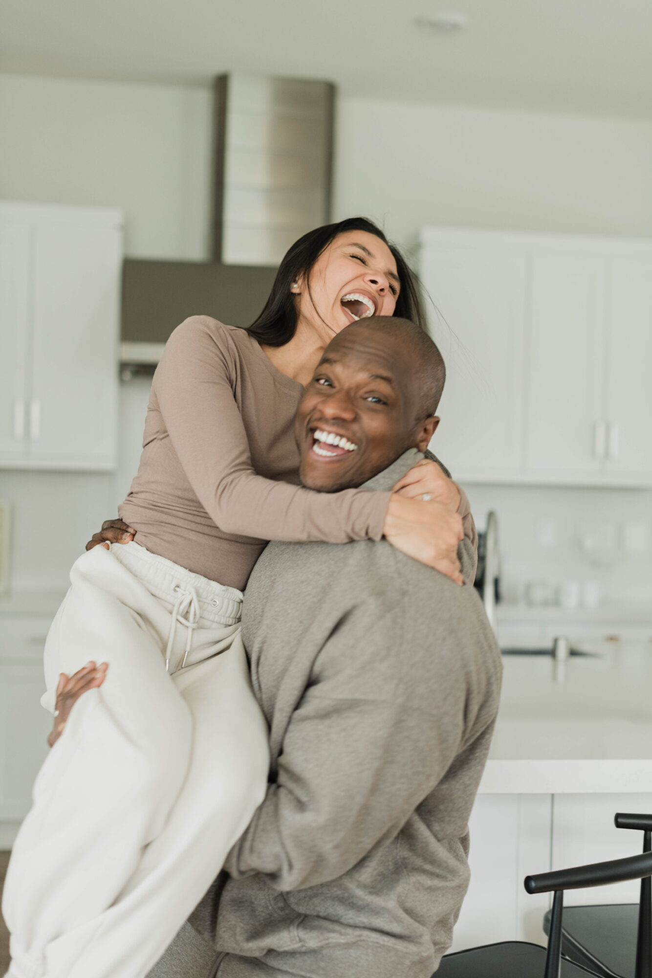Two people hugging and laughing in a bright kitchen, one sitting and the other standing on their lap.
