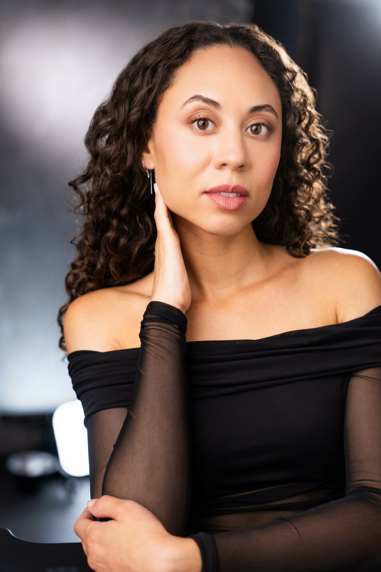 Woman with curly hair wearing an off-shoulder black top, touching her face with her right hand, looking at the camera.