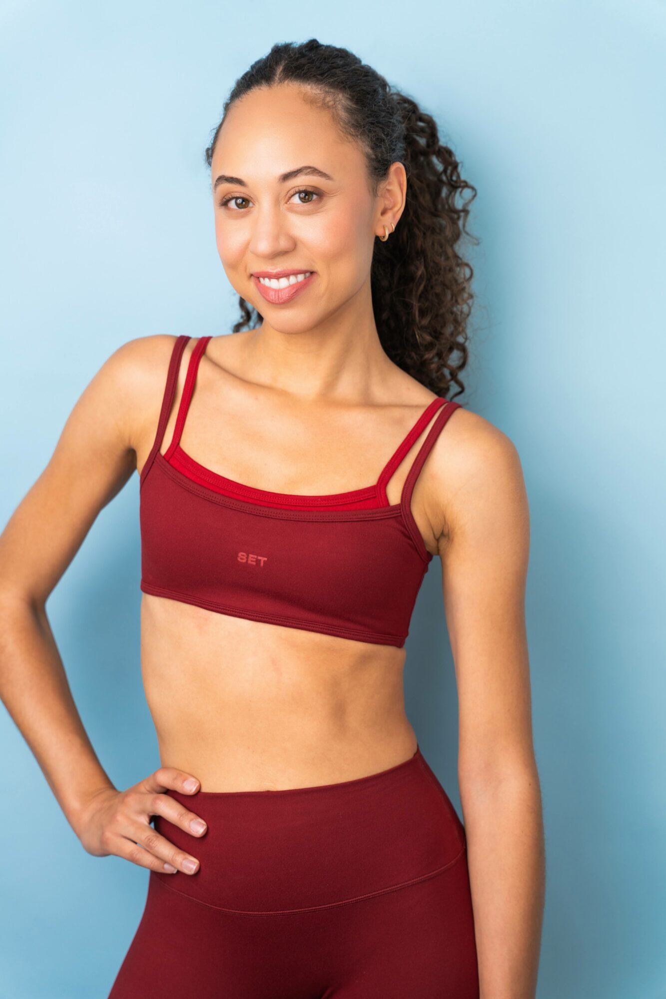 Young woman with curly hair in a red sports bra and matching leggings, smiling, standing against a light blue background.