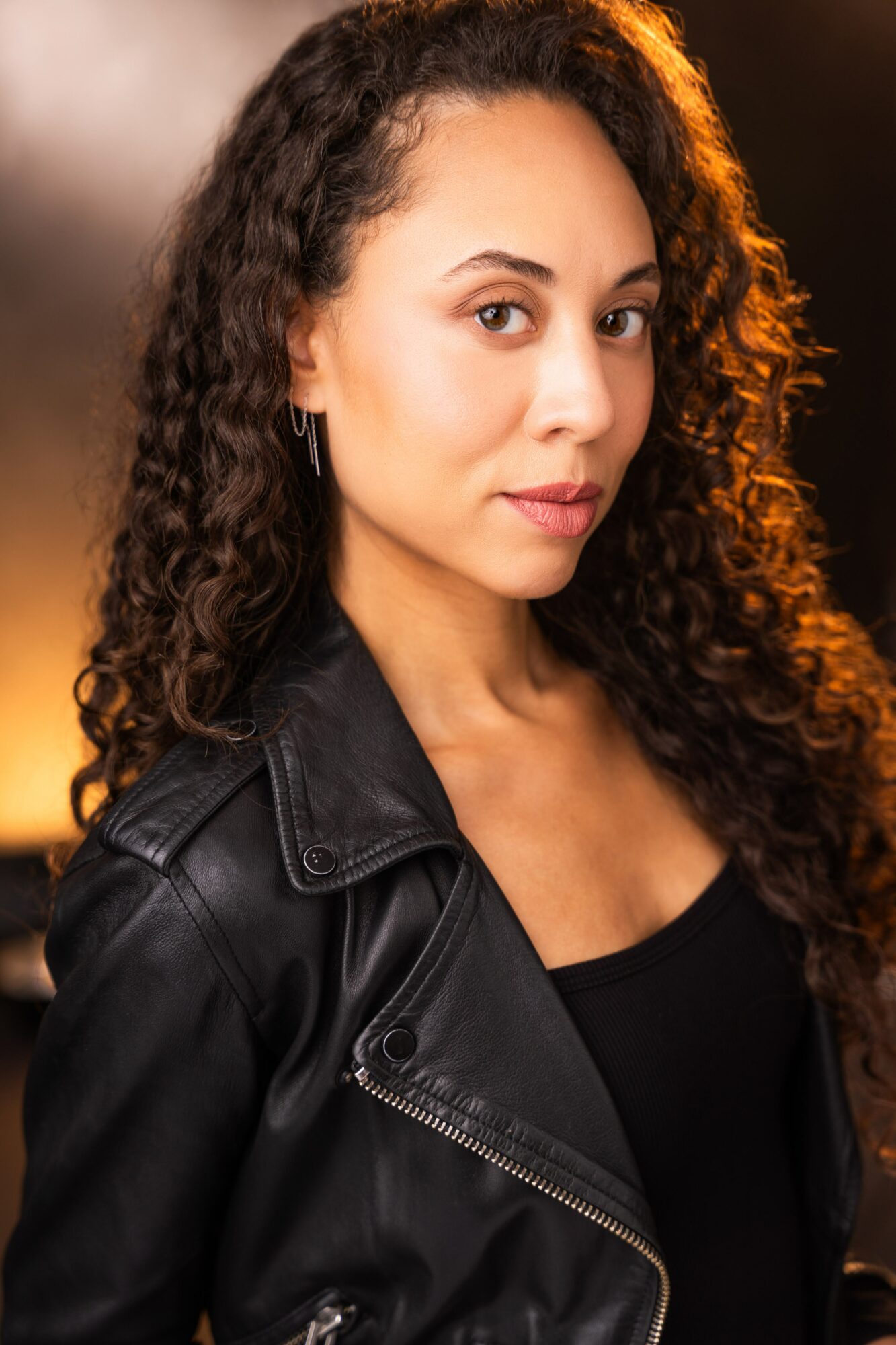 Portrait of a woman with curly hair wearing a black leather jacket, looking at the camera with a neutral expression.