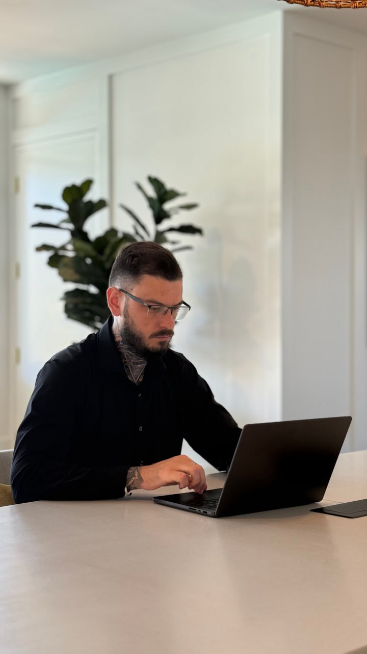 Man with glasses working on a laptop at a table, with plants and white walls in the background.