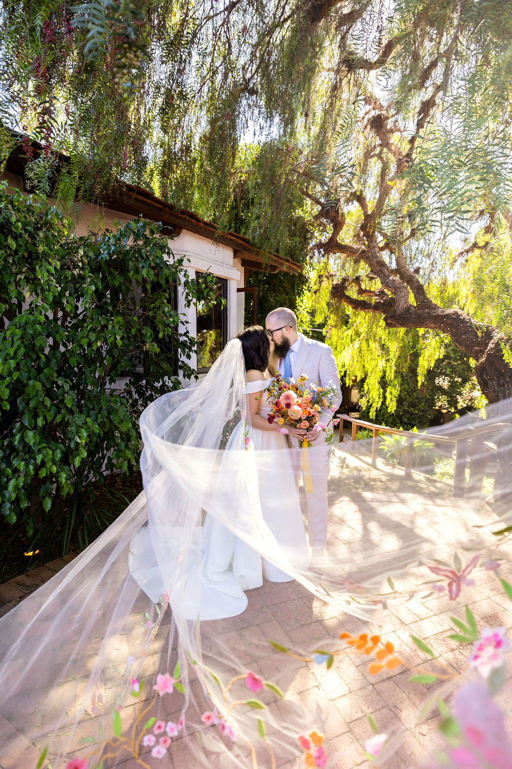 Bride and groom standing outdoors with trees and sunlight, bride holding a bouquet, veil flowing, wedding attire, garden setting.
