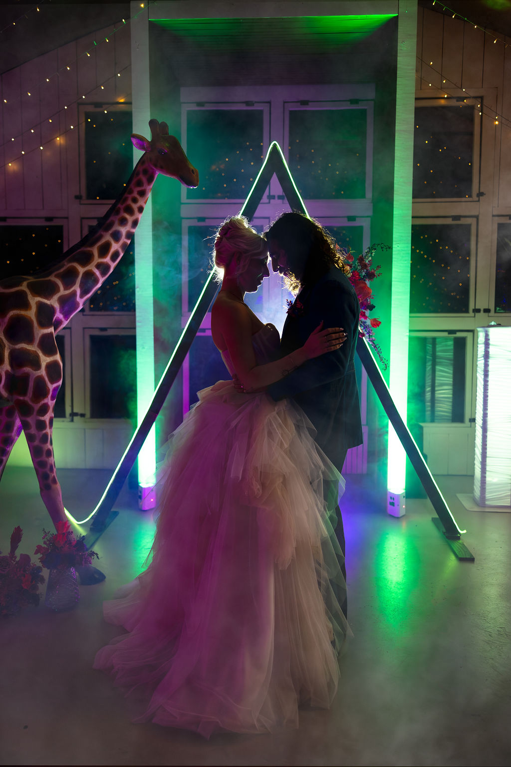 Two people in formal attire embrace in front of a geometric light installation, with a giraffe figure nearby, in a decorated venue.