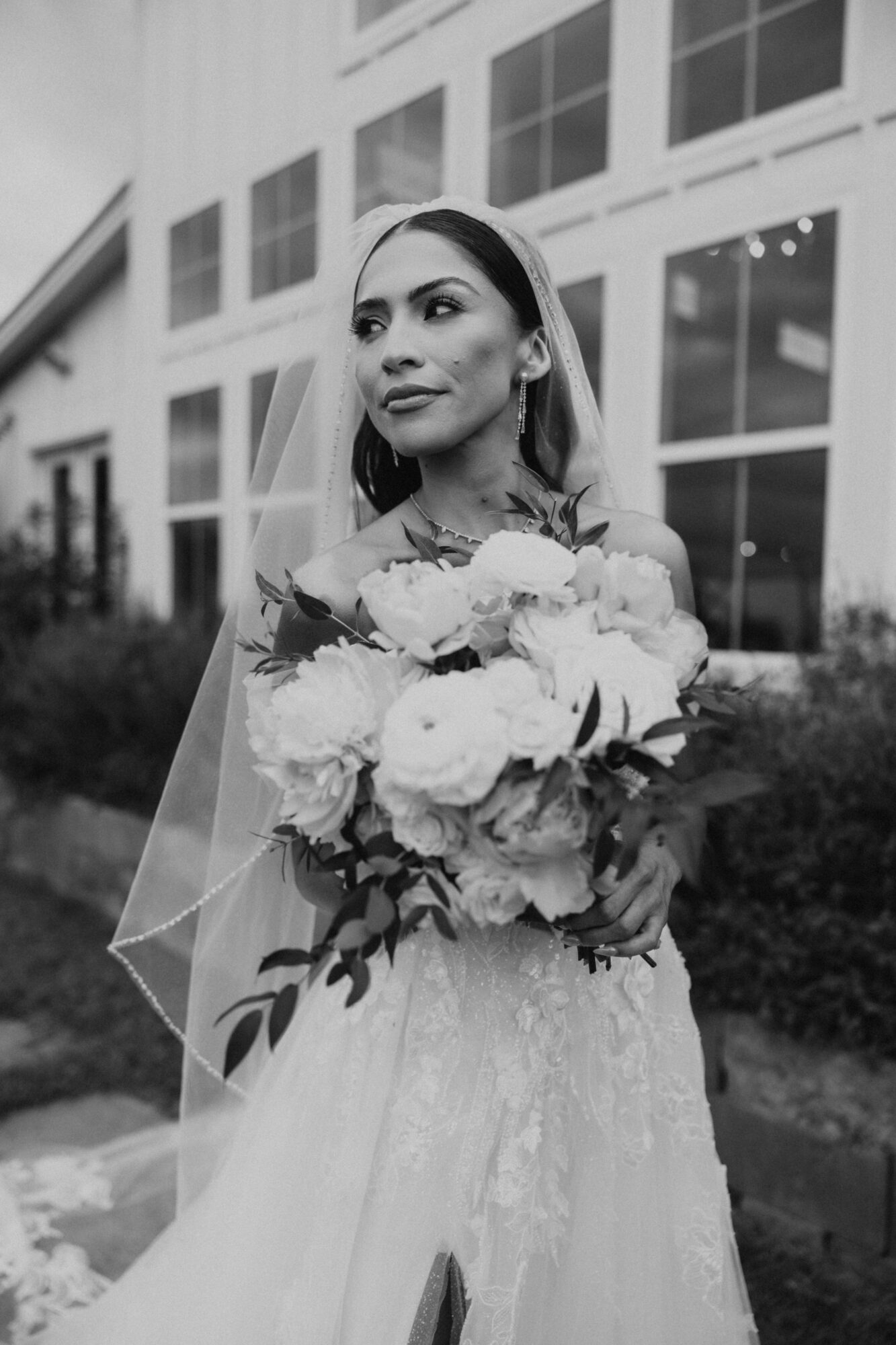 Woman in wedding dress holding a bouquet of flowers outdoors, with modern building in background.